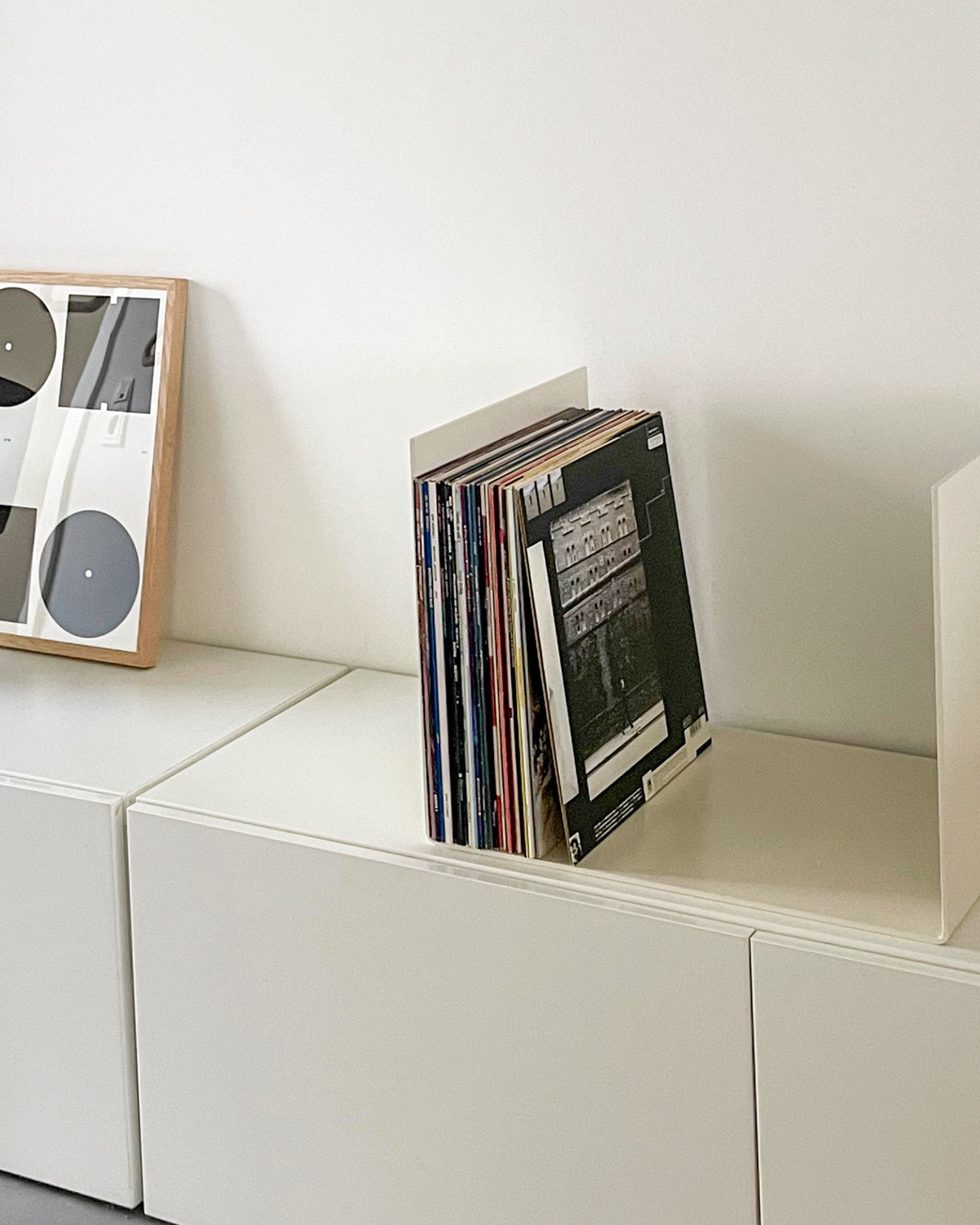 Nahaufnahme einer cremeweißen Metallablage auf einer weißen Kommode, dekoriert mit Vinyl-Schallplatten und einem gerahmten Kunstobjekt, modernes Wohnaccessoire