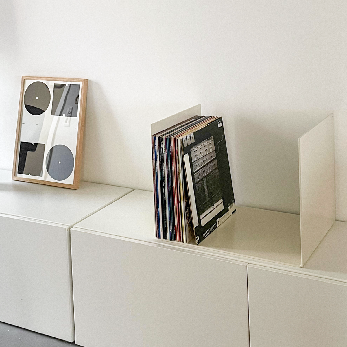 Nahaufnahme einer cremeweißen Metallablage auf einer weißen Kommode, dekoriert mit Vinyl-Schallplatten und einem gerahmten Kunstobjekt, modernes Wohnaccessoire
