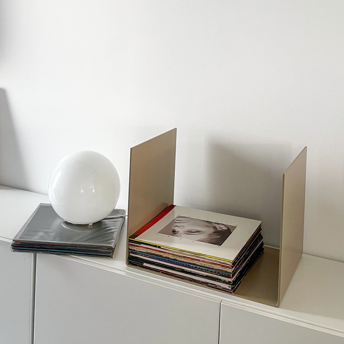 Nahaufnahme einer graubeigen Metallablage auf einer weißen Kommode, dekoriert mit Vinyl-Schallplatten und einem gerahmten Kunstobjekt, modernes Wohnaccessoire