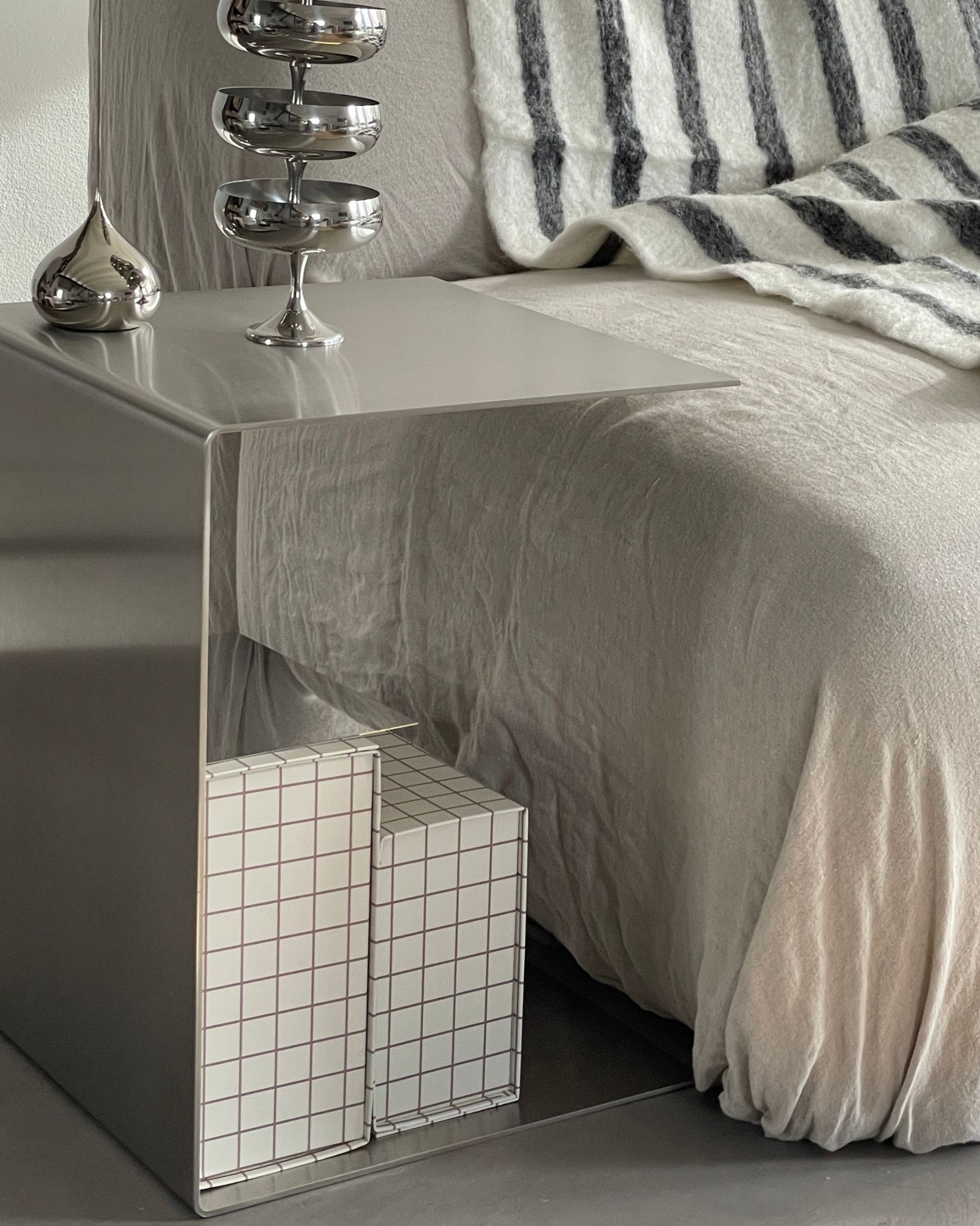 Gebürsteter Edelstahl-Beistelltisch neben einem Sofa, dekoriert mit Edelstahl-Accessoires und gestapelten Boxen mit Rastermuster