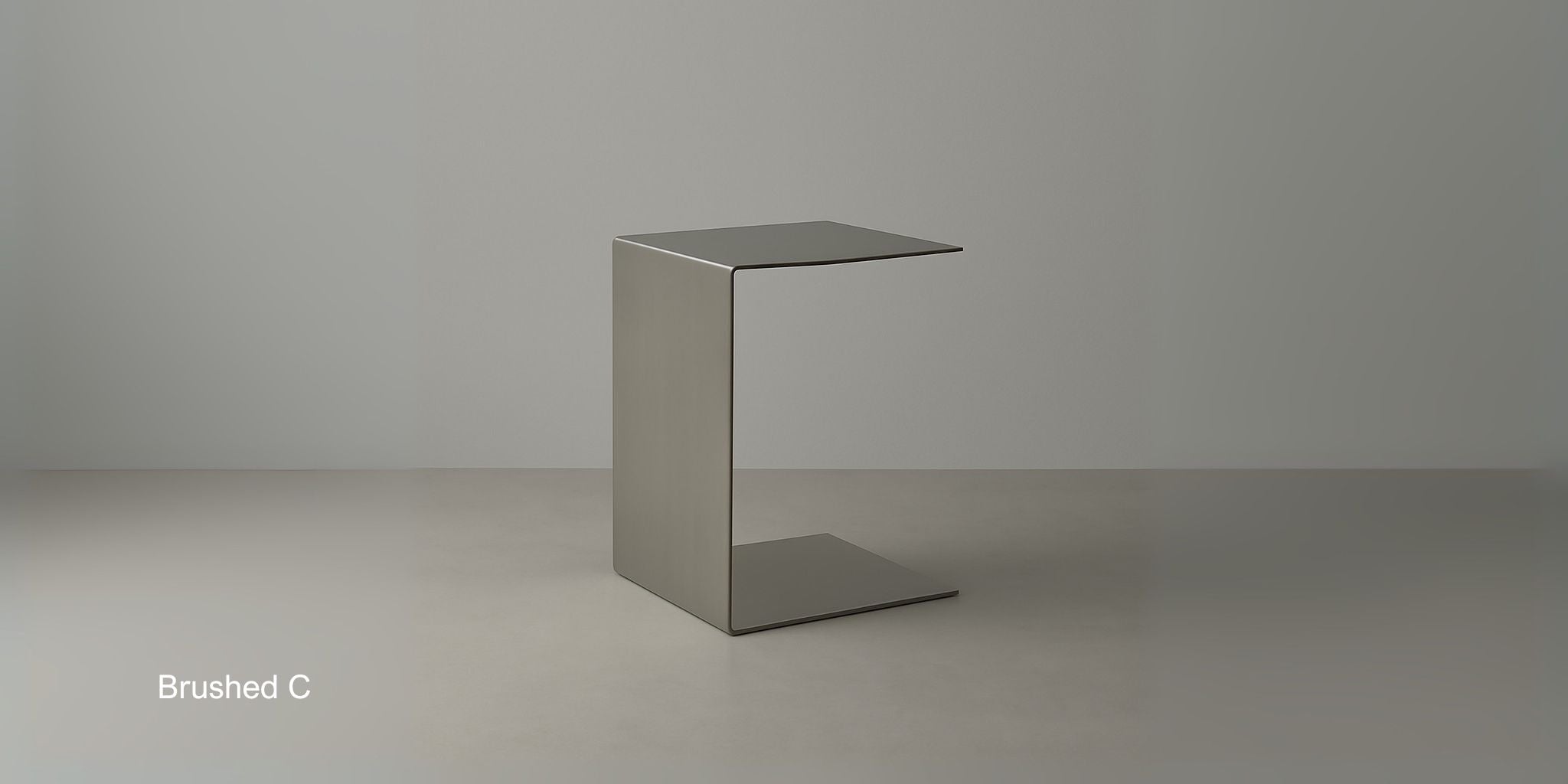 Beistelltisch Brushed C aus gebürstetem Edelstahl im minimalistischen C-Design auf hellem Boden vor grauer Wand