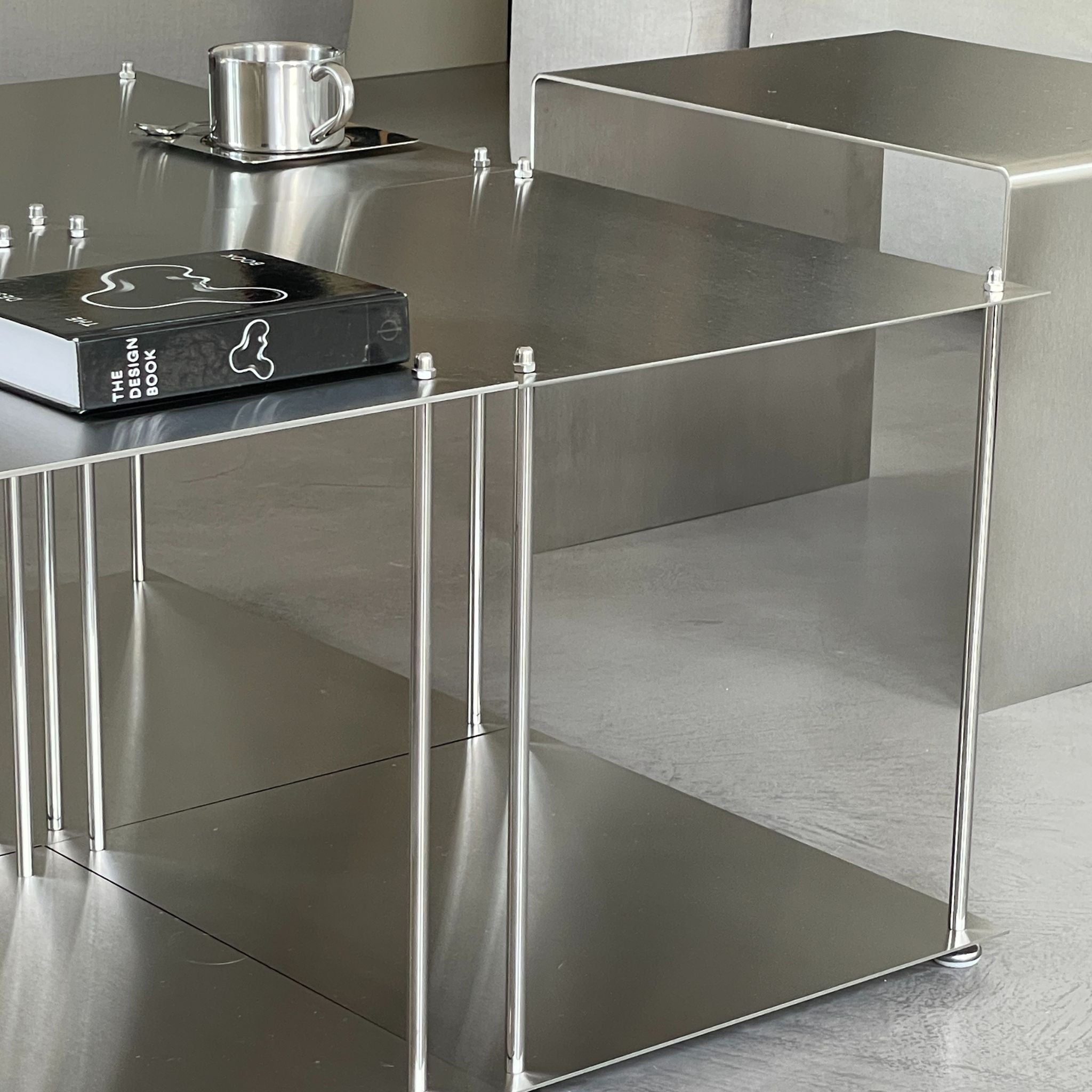Nahaufnahme eines quadratischen Modulum Couchtisches aus gebürstetem Edelstahl mit zwei Ebenen und sichtbaren Verschraubungen auf glattem Boden im modernen Wohnraum