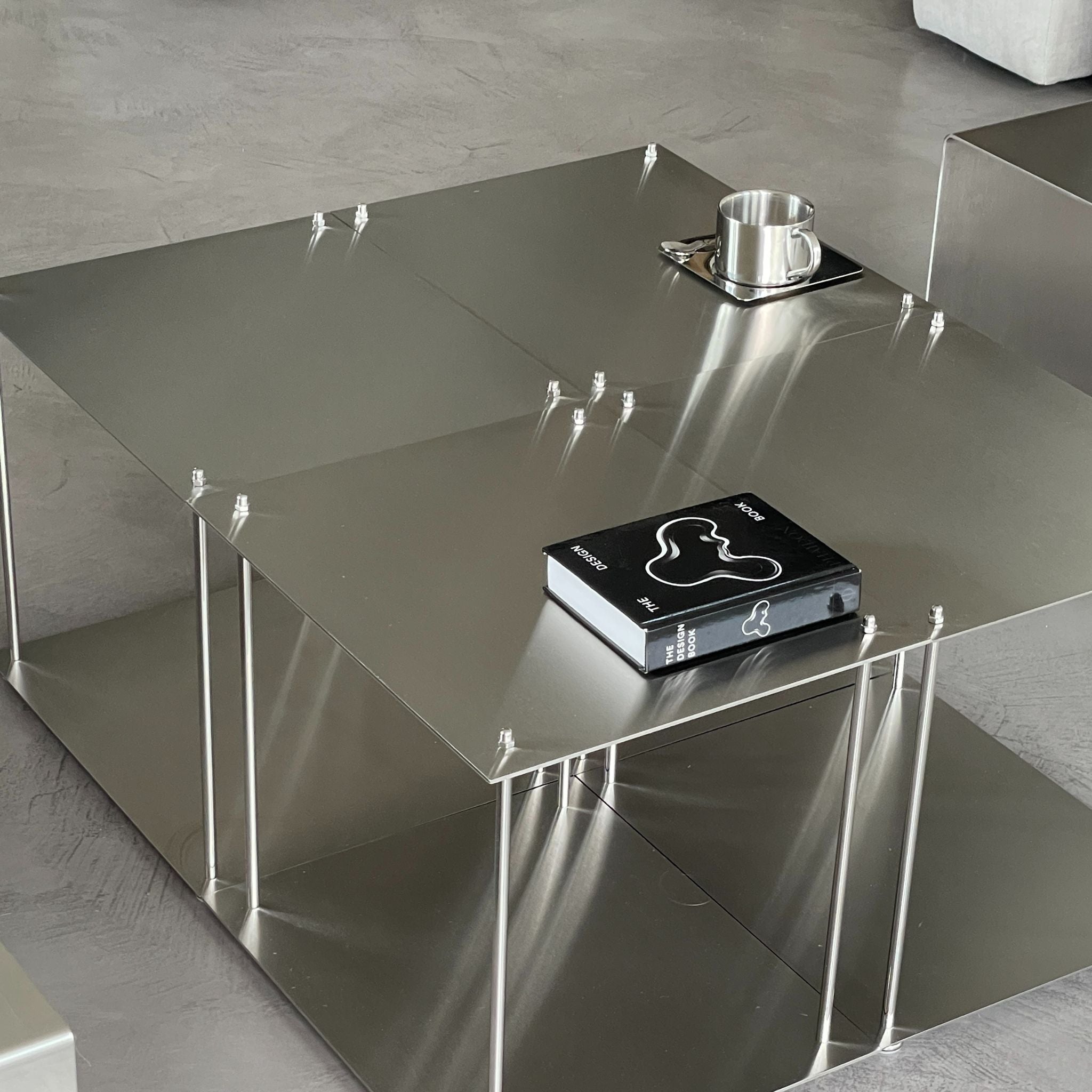 Quadratischer Modulum Couchtisch aus gebürstetem Edelstahl mit zwei Ebenen, darauf ein Buch und eine Edelstahl-Tasse auf Untersetzer, stehend auf glattem Boden in modernem Wohnraum