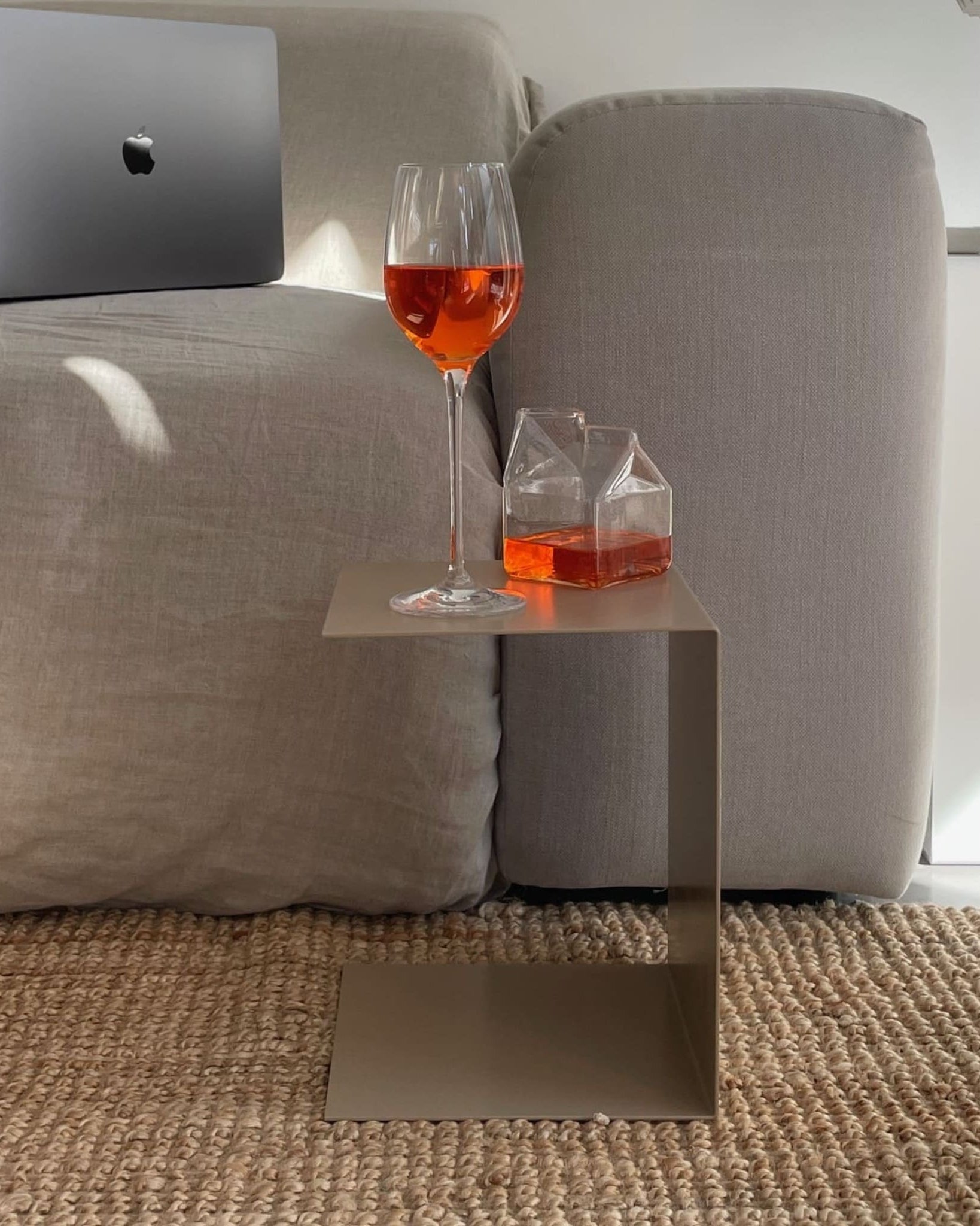 Kleiner graubeiger Beistelltisch mit Aperol-Glas und Karaffe, neben grauem Sofa auf geflochtenem Teppich