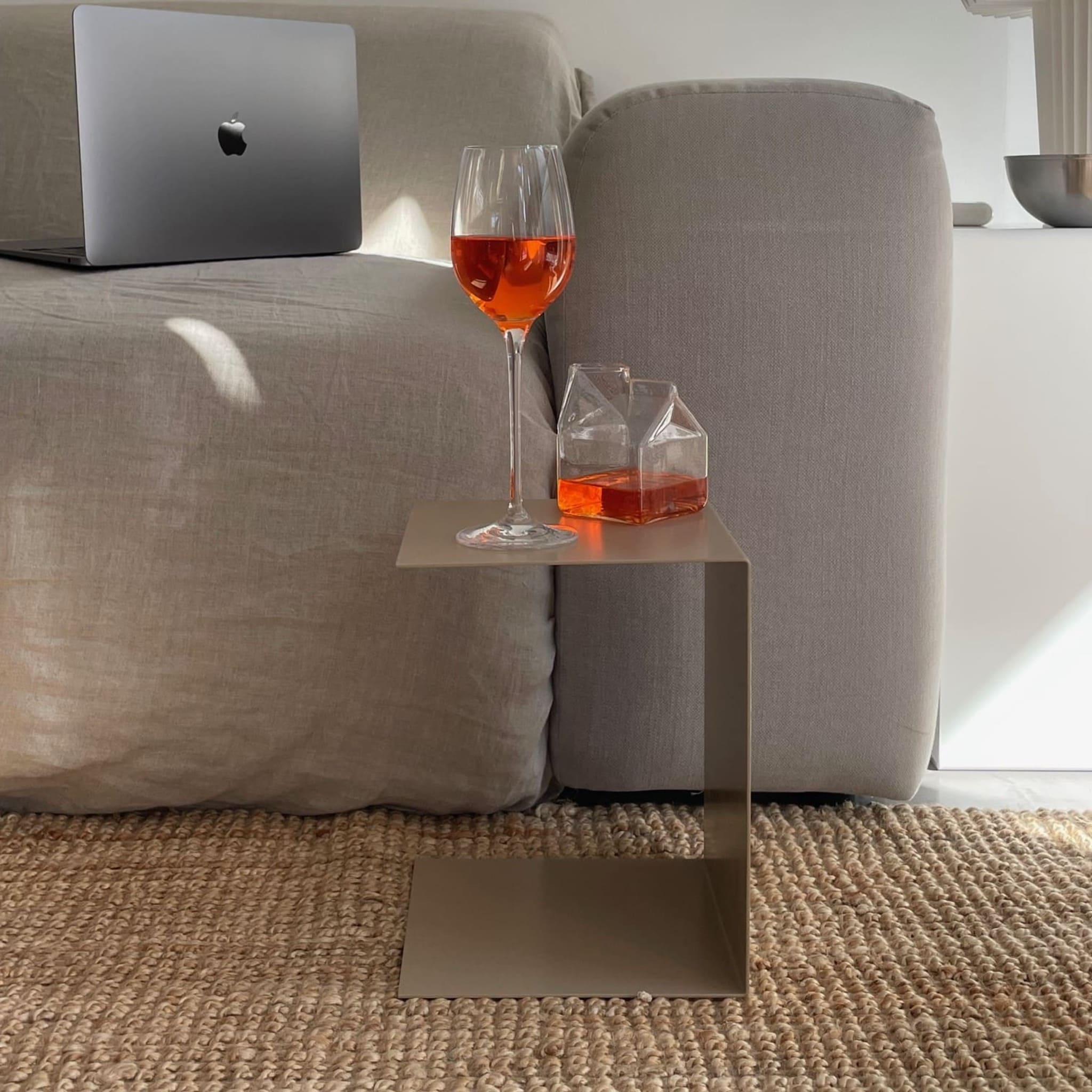 Kleiner graubeiger Beistelltisch mit Aperol-Glas und Karaffe, neben grauem Sofa auf geflochtenem Teppich