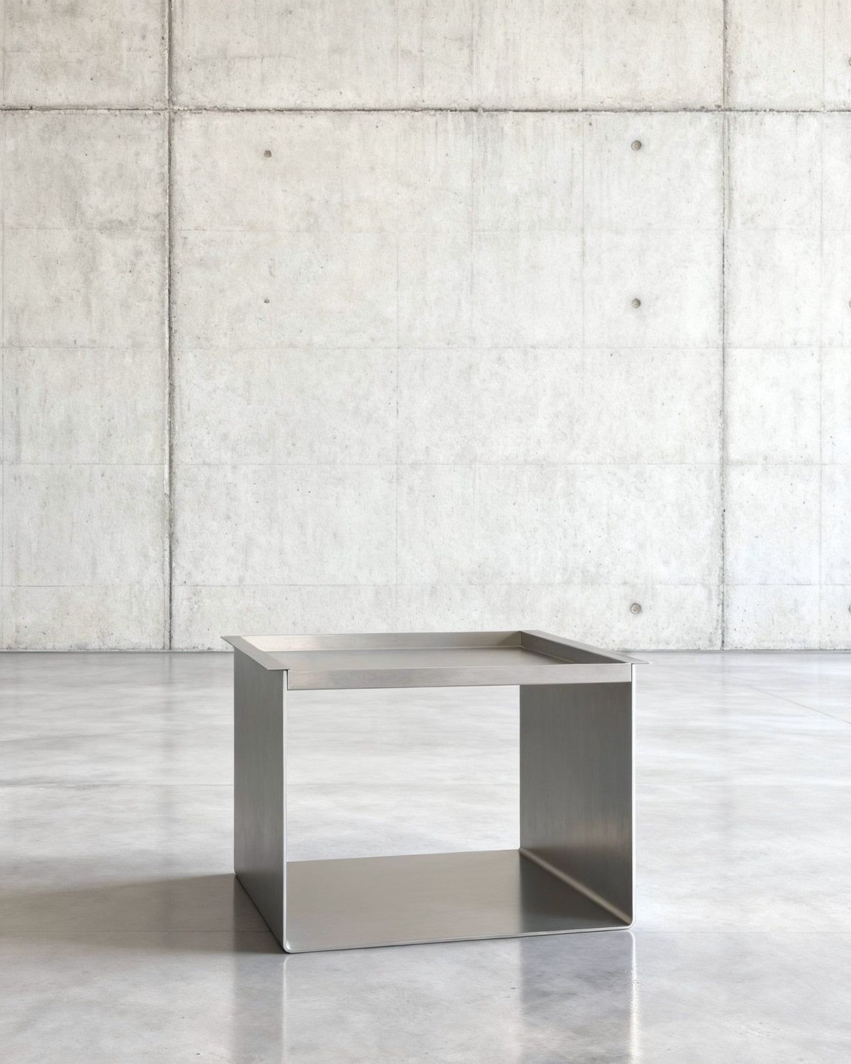 Tray Table Brushed C Beistelltisch in gebürstetem Edelsthahl auf grauen Betonboden vor grauer Betonwand mit Löchern