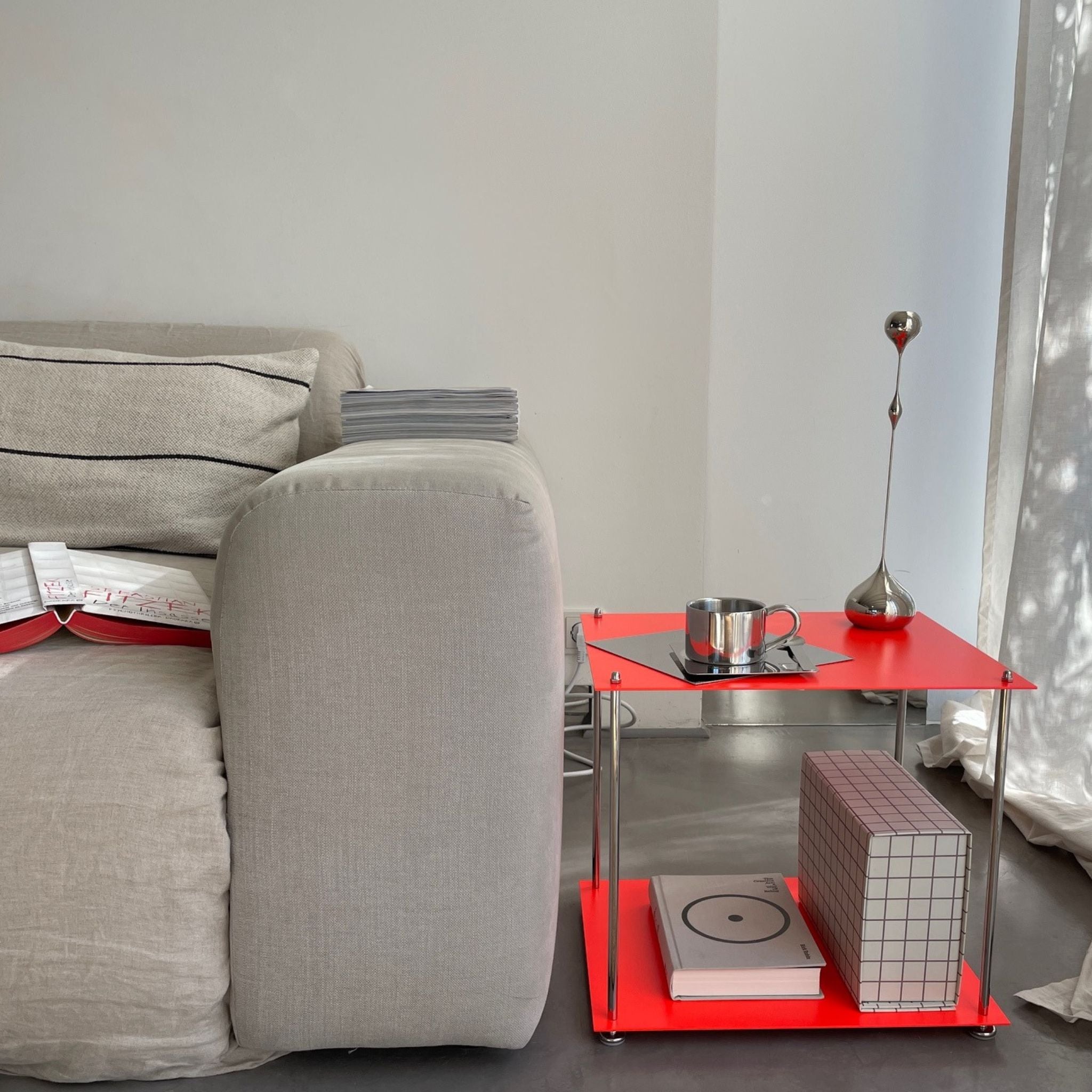 Leuchtroter Beistelltisch aus Edelstahl neben hellem Sofa, dekoriert mit Designobjekten und Büchern