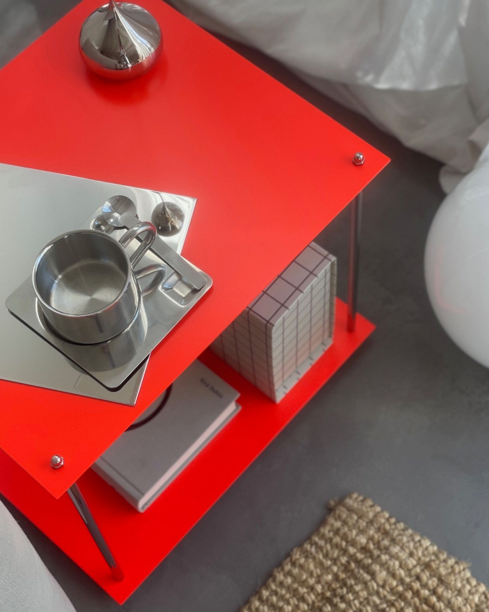 Leuchtroter Design-Beistelltisch mit Edelstahlgestell, Spiegelplatte, Tasse und Deko neben Sofa auf grauem Boden