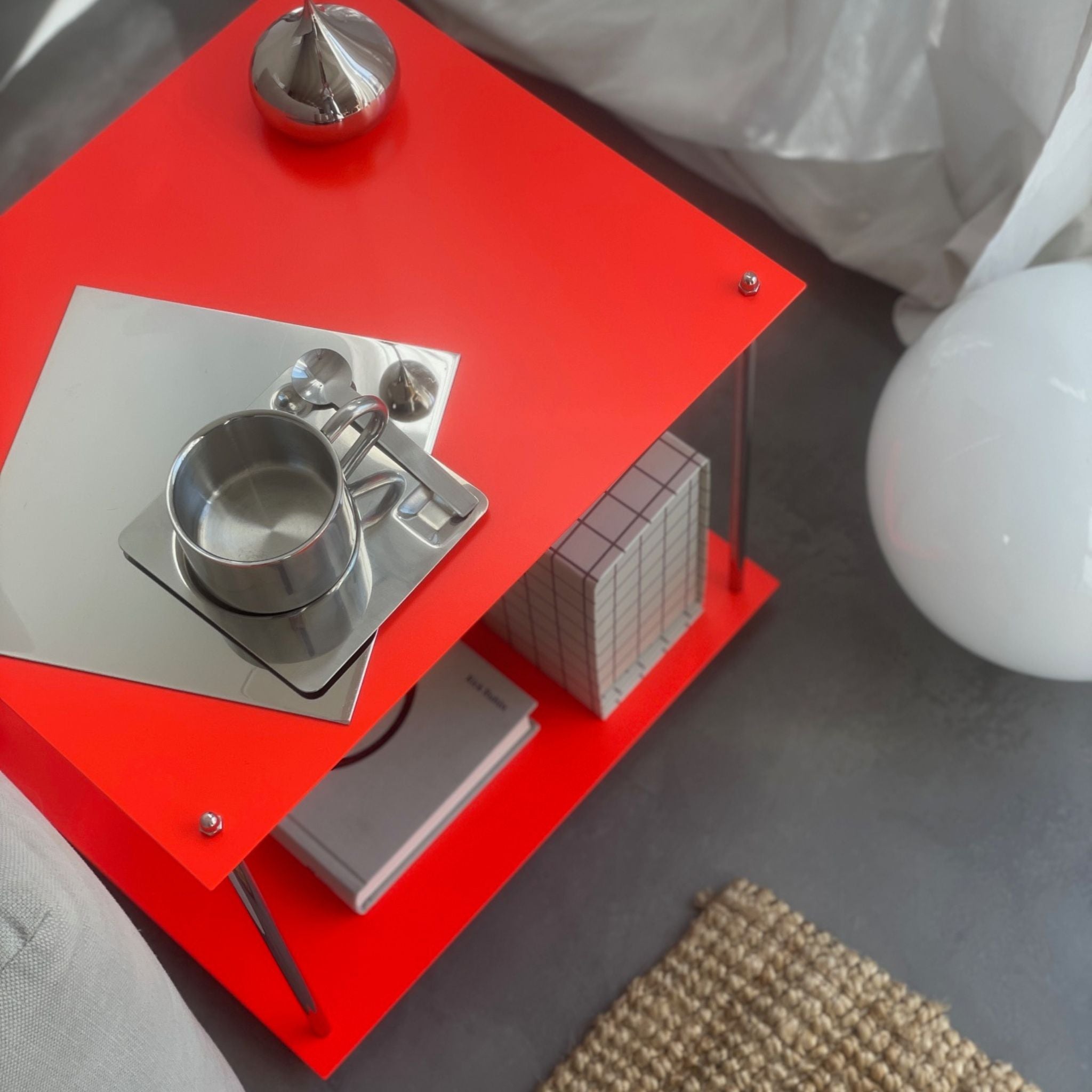 Leuchtroter Design-Beistelltisch mit Edelstahlgestell, Spiegelplatte, Tasse und Deko neben Sofa auf grauem Boden