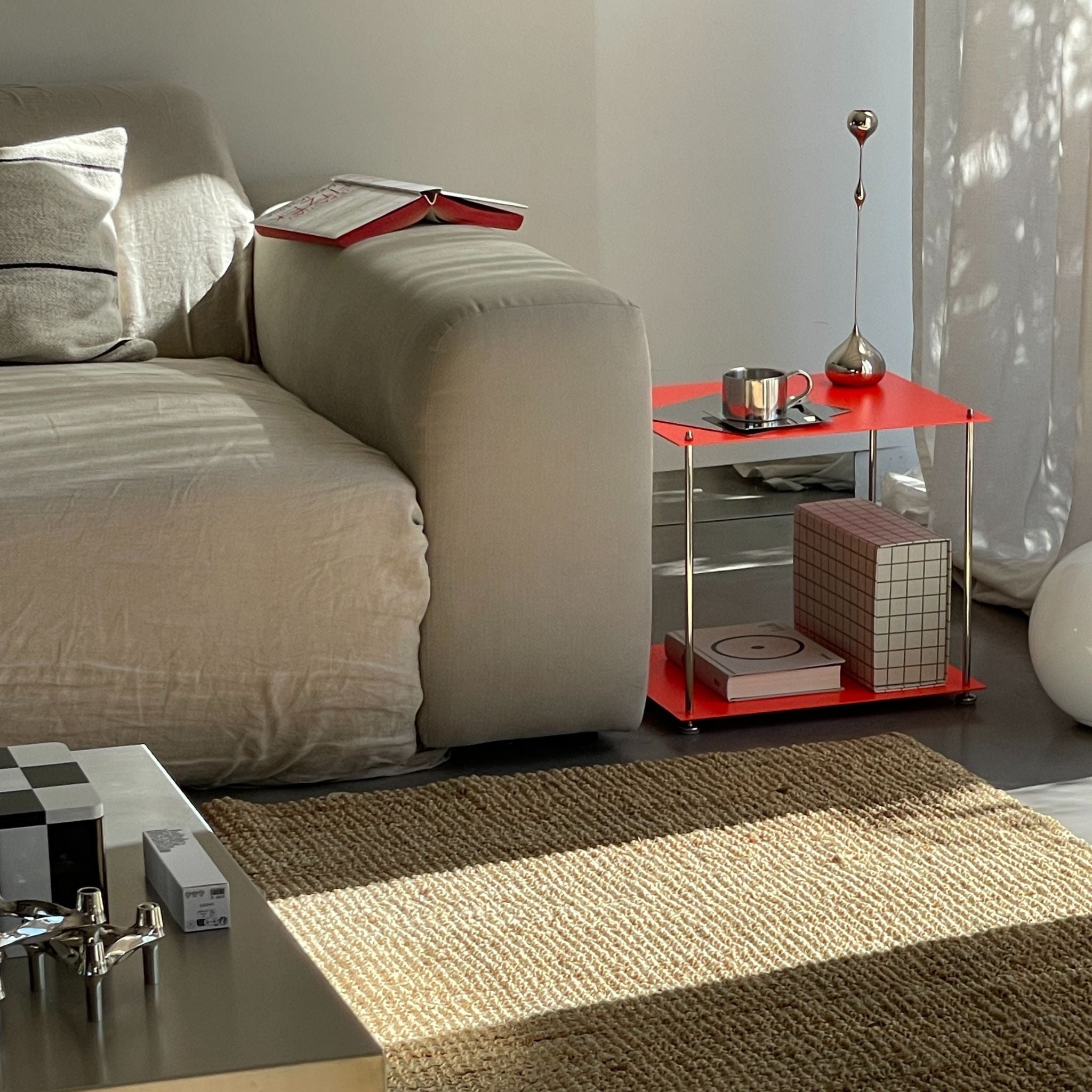 Modernes Wohnzimmer mit leuchtrotem Beistelltisch aus Edelstahl, neben Sofa, Bücher und Deko im Sonnenlicht
