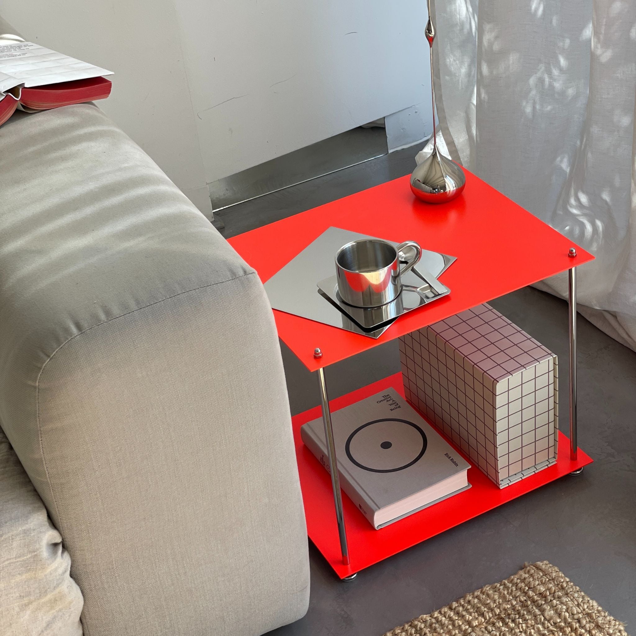 Leuchtroter Beistelltisch aus Edelstahl neben Sofa mit Tasse, Dekoobjekten und Designbuch auf grauem Boden
