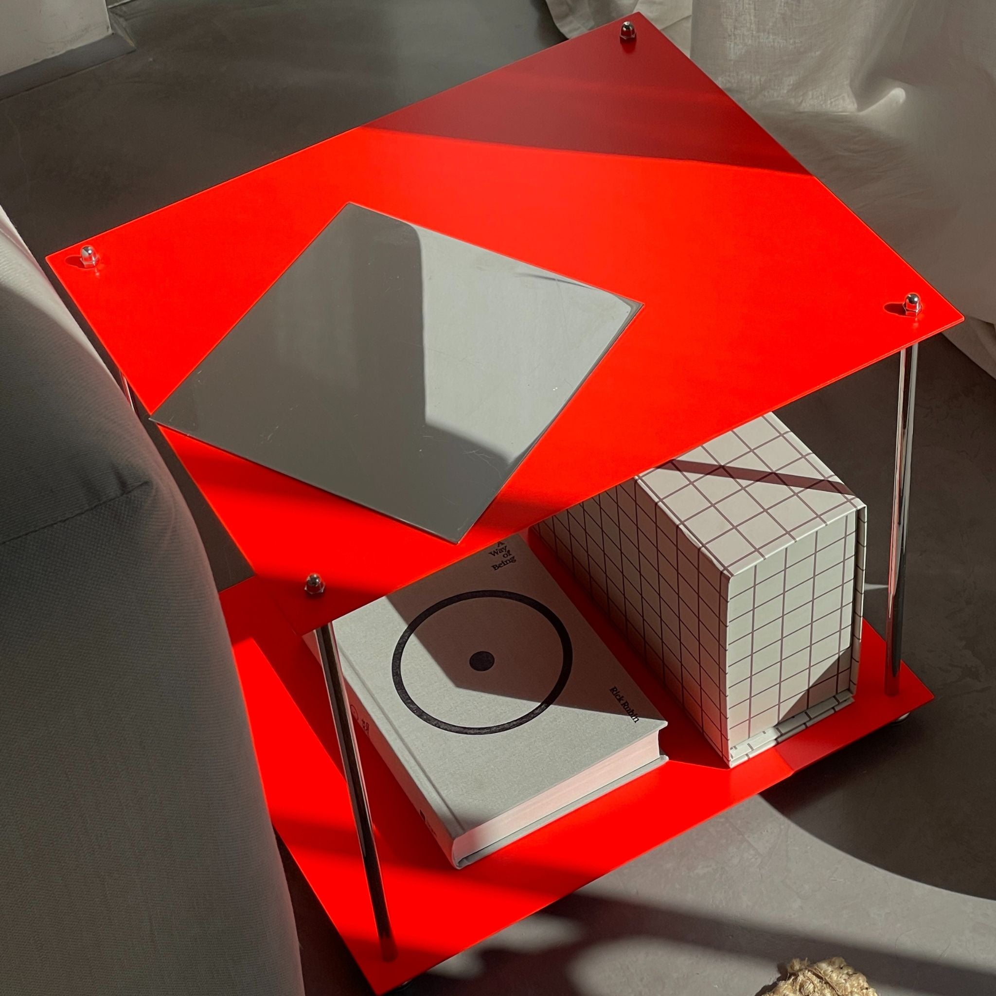 Detailansicht eines leuchtroten Beistelltischs mit Designbuch, Spiegelplatte und Rasterbox auf grauem Boden