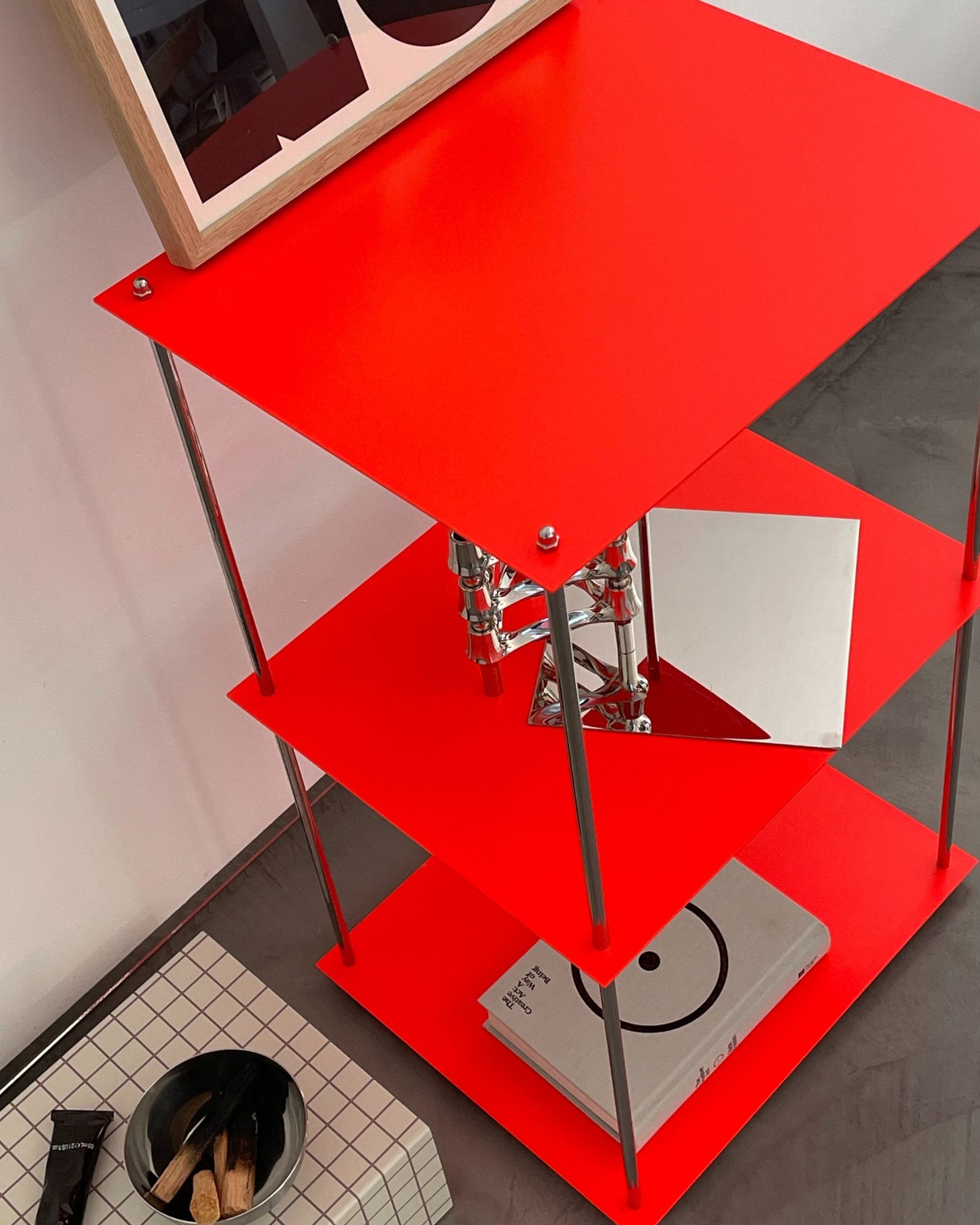 Modulares Designregal MODULUM in Leuchtrot mit drei Böden, dekoriert mit Buch und Kerzenhalter, vor weißer Wand auf Betonboden