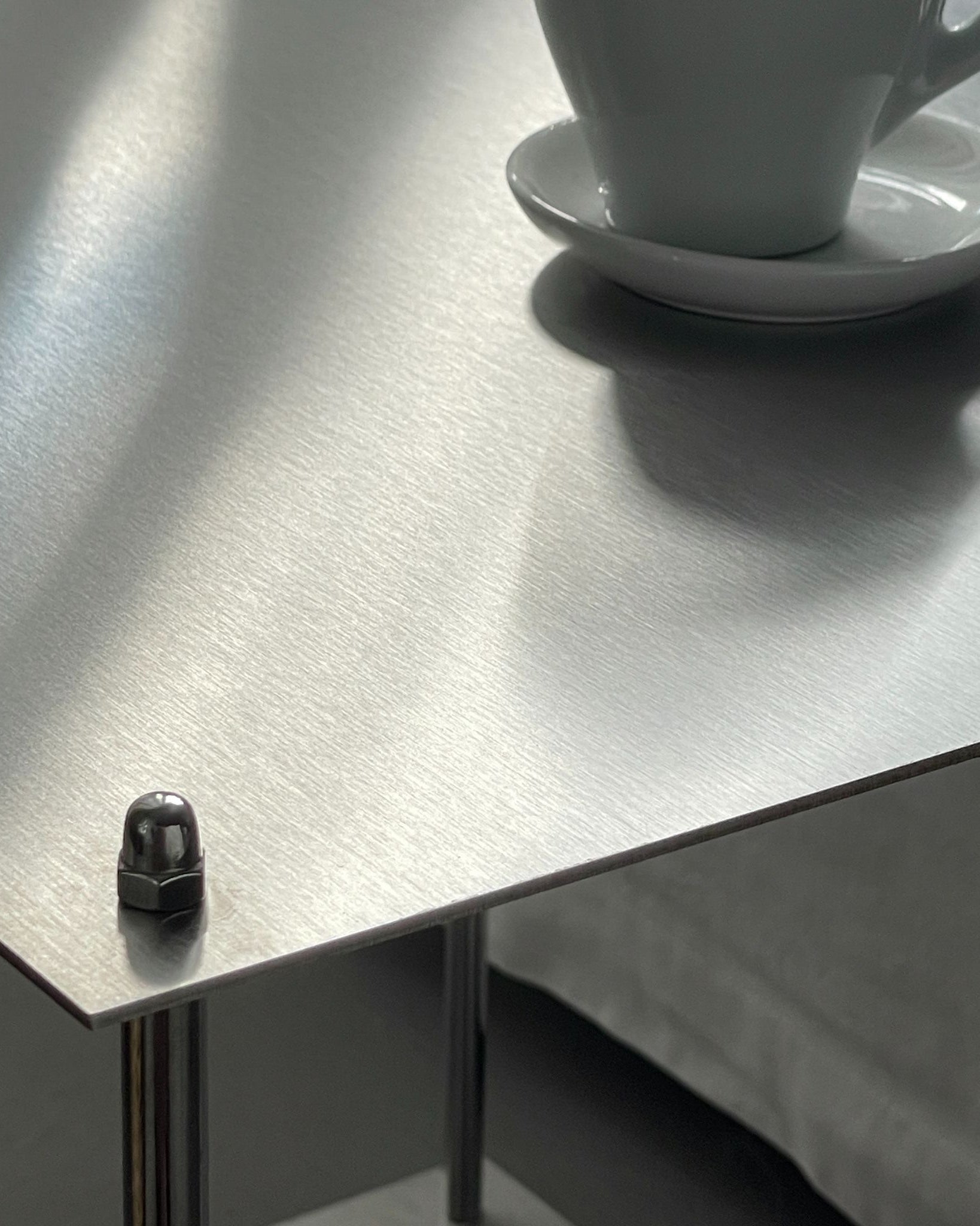 Nahaufnahme der gebürsteten Edelstahloberfläche eines Modulum Beistelltisches, Detail der Schraubverbindung und einer Kaffeetasse.