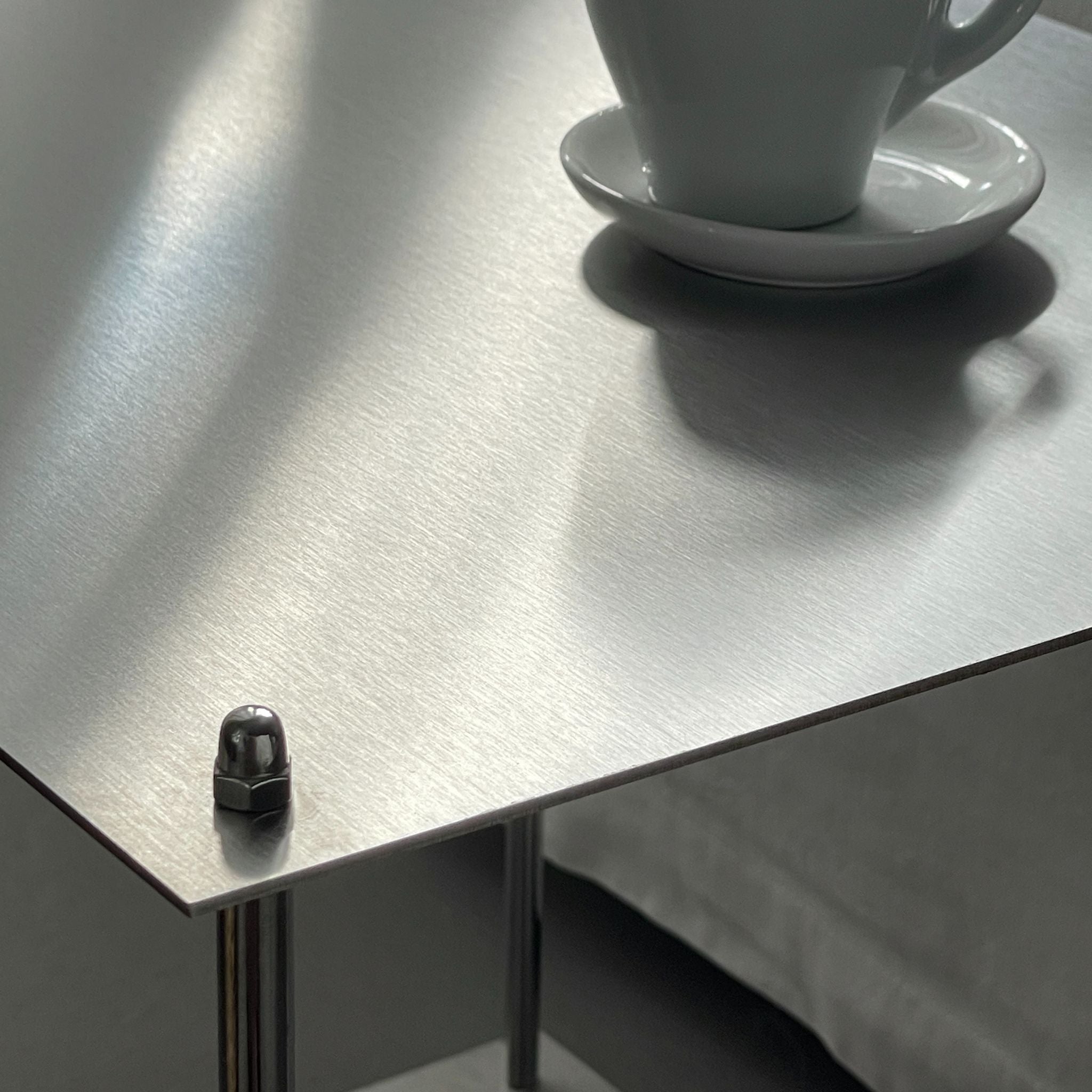 Nahaufnahme der gebürsteten Edelstahloberfläche eines Modulum Beistelltisches, Detail der Schraubverbindung und einer Kaffeetasse.