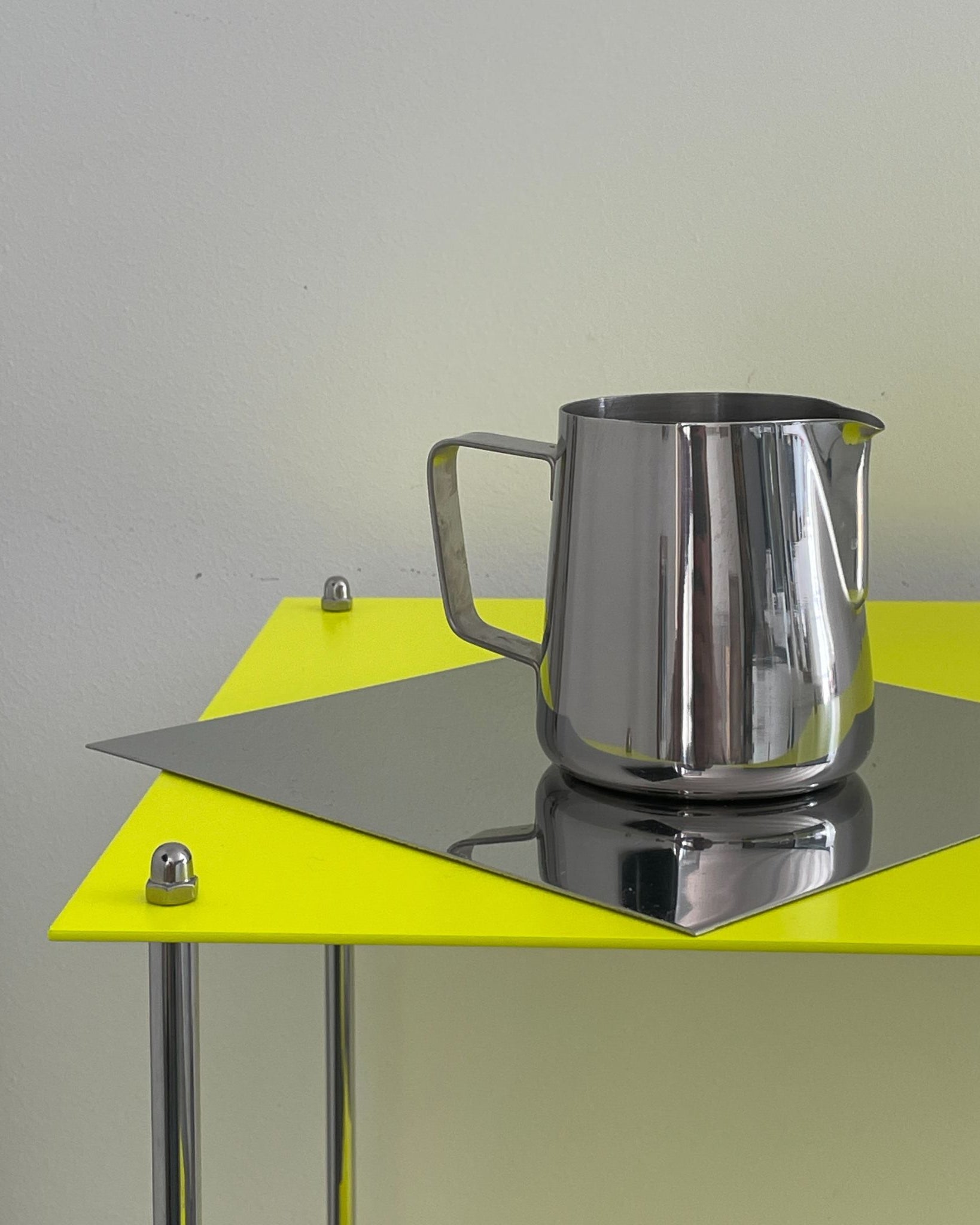 Edelstahlkanne auf spiegelnder Platte eines leuchtgelben MODULUM Regals, modernes Designobjekt vor heller Wand