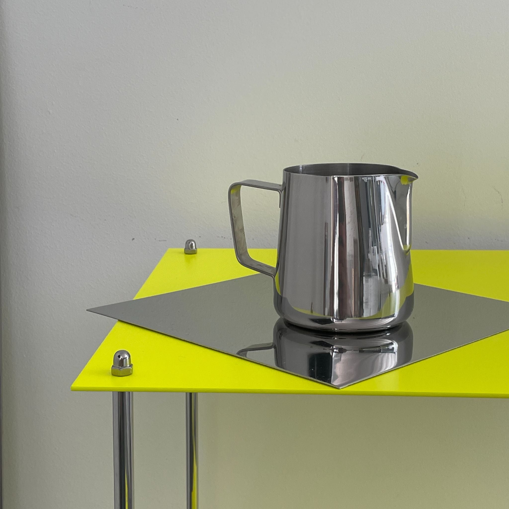 Edelstahlkanne auf spiegelnder Platte eines leuchtgelben MODULUM Regals, modernes Designobjekt vor heller Wand