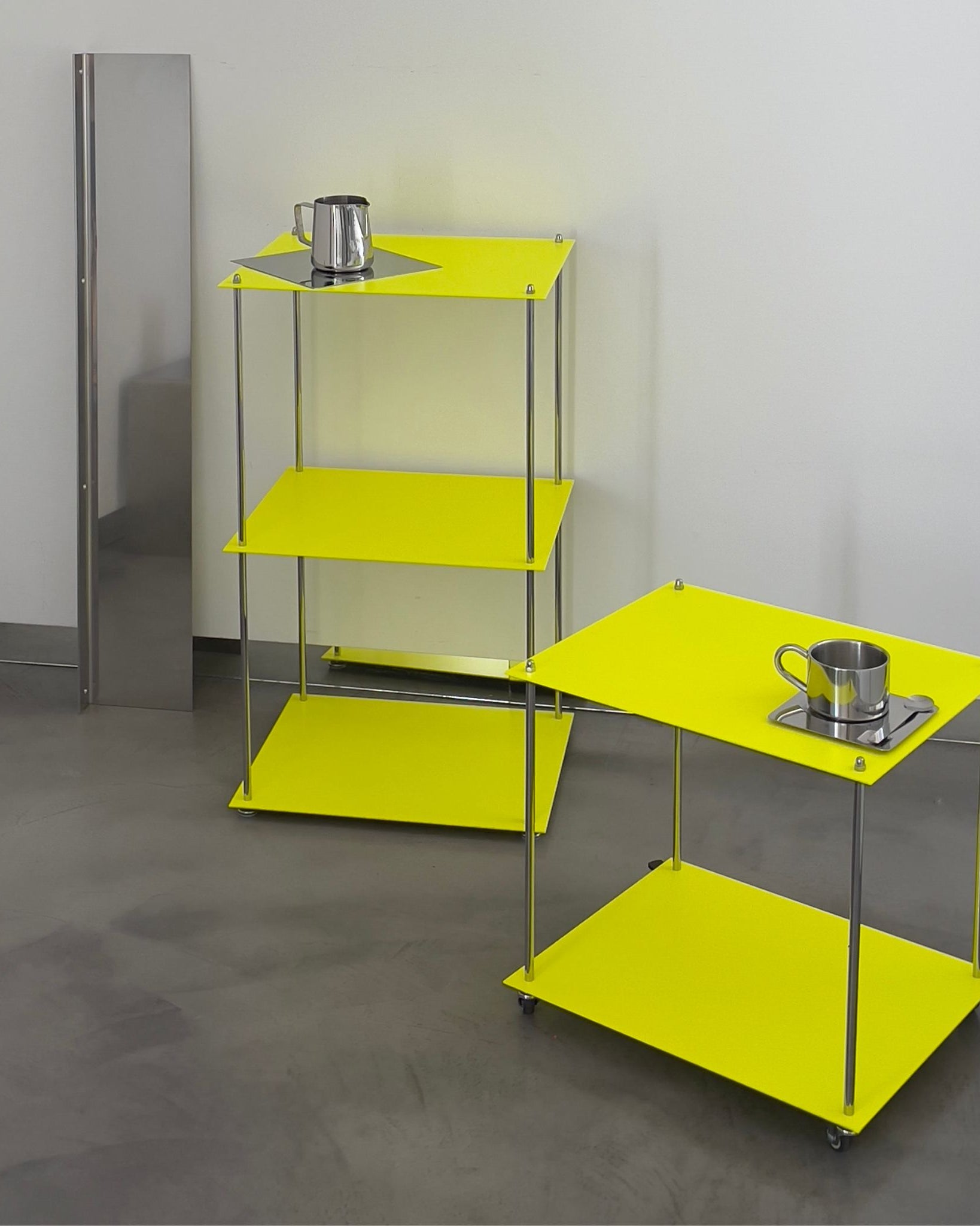 Zwei leuchtgelbe MODULUM Regale mit Edelstahlkanne und Tasse, eines davon mobil auf Rollen, vor grauer Wand und Boden
