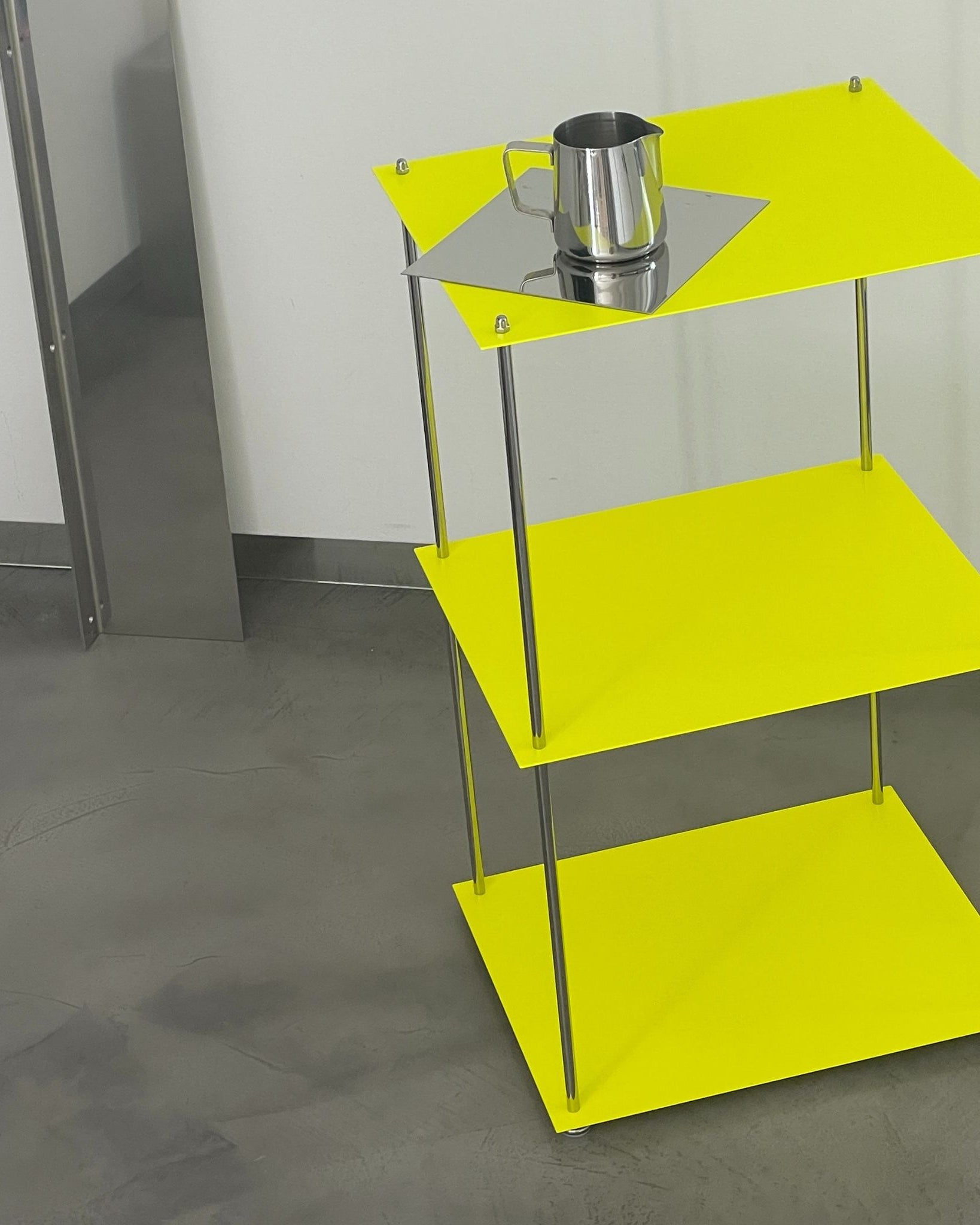 Leuchtgelbes MODULUM Regal mit Edelstahlkanne auf quadratischer Spiegelplatte vor grauem Betonboden und Wand