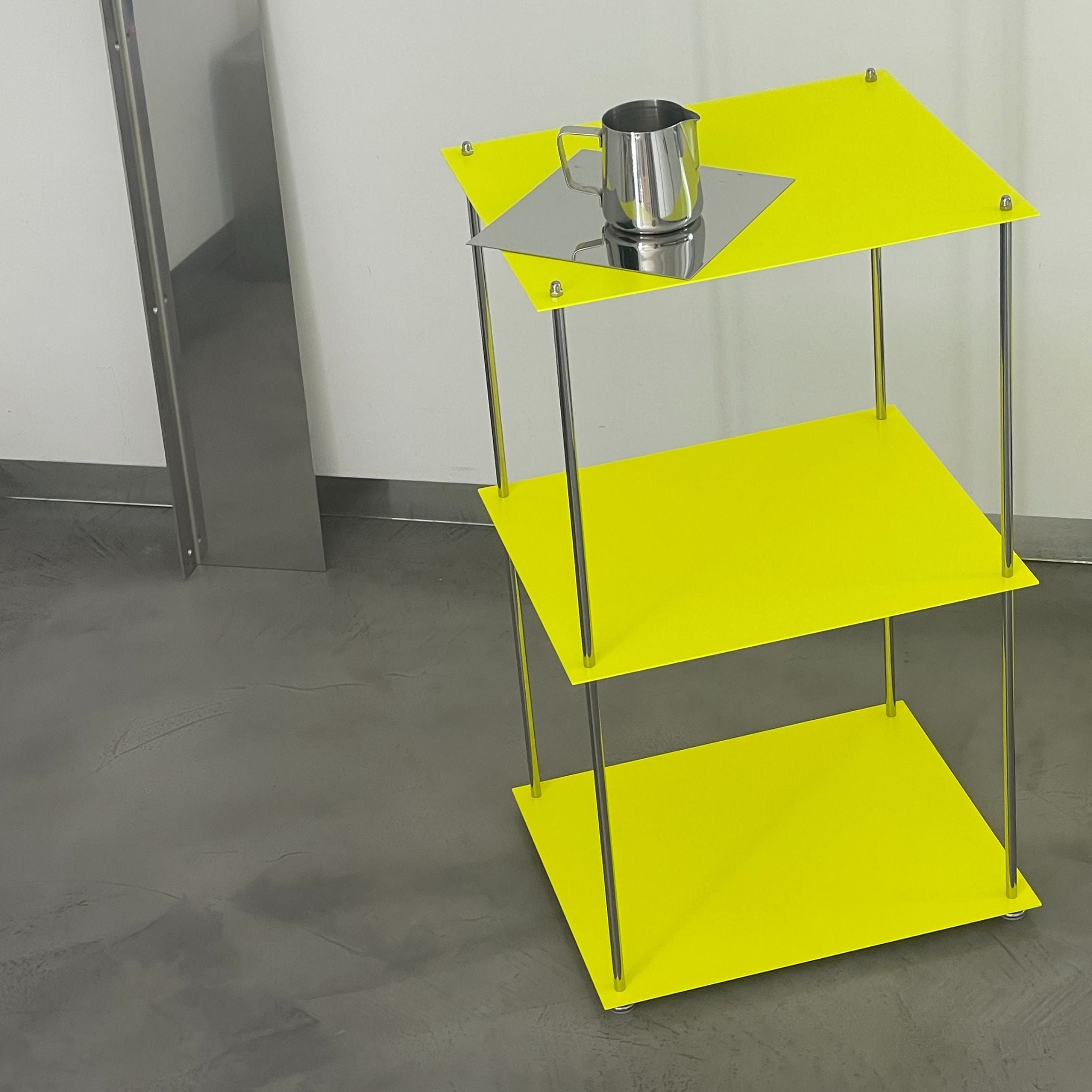 Leuchtgelbes MODULUM Regal mit Edelstahlkanne auf quadratischer Spiegelplatte vor grauem Betonboden und Wand