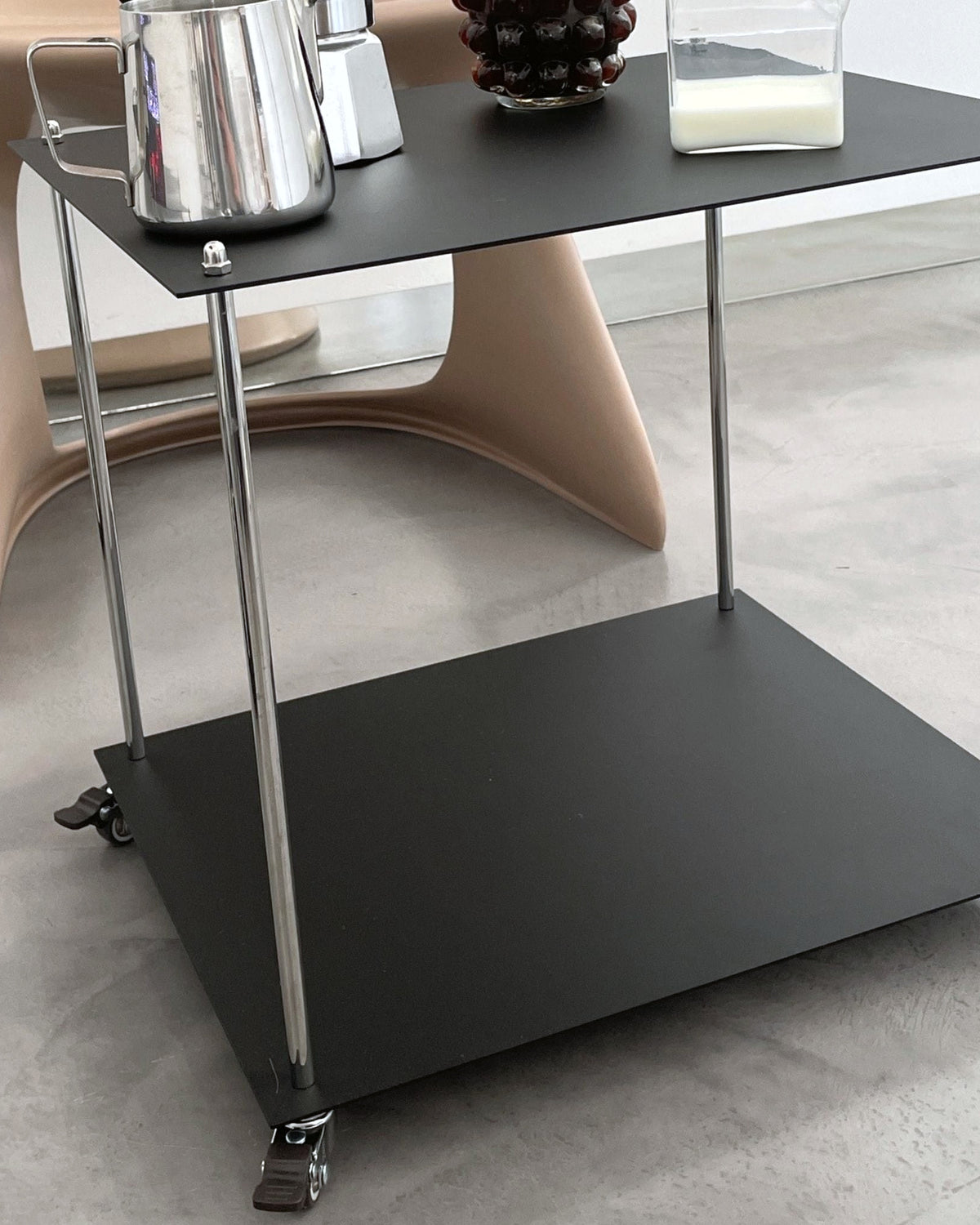 Moderner Servierwagen aus schwarzem Stahl mit Rollen im minimalistischen Interior neben Designstuhl und Kaffeegeschirr