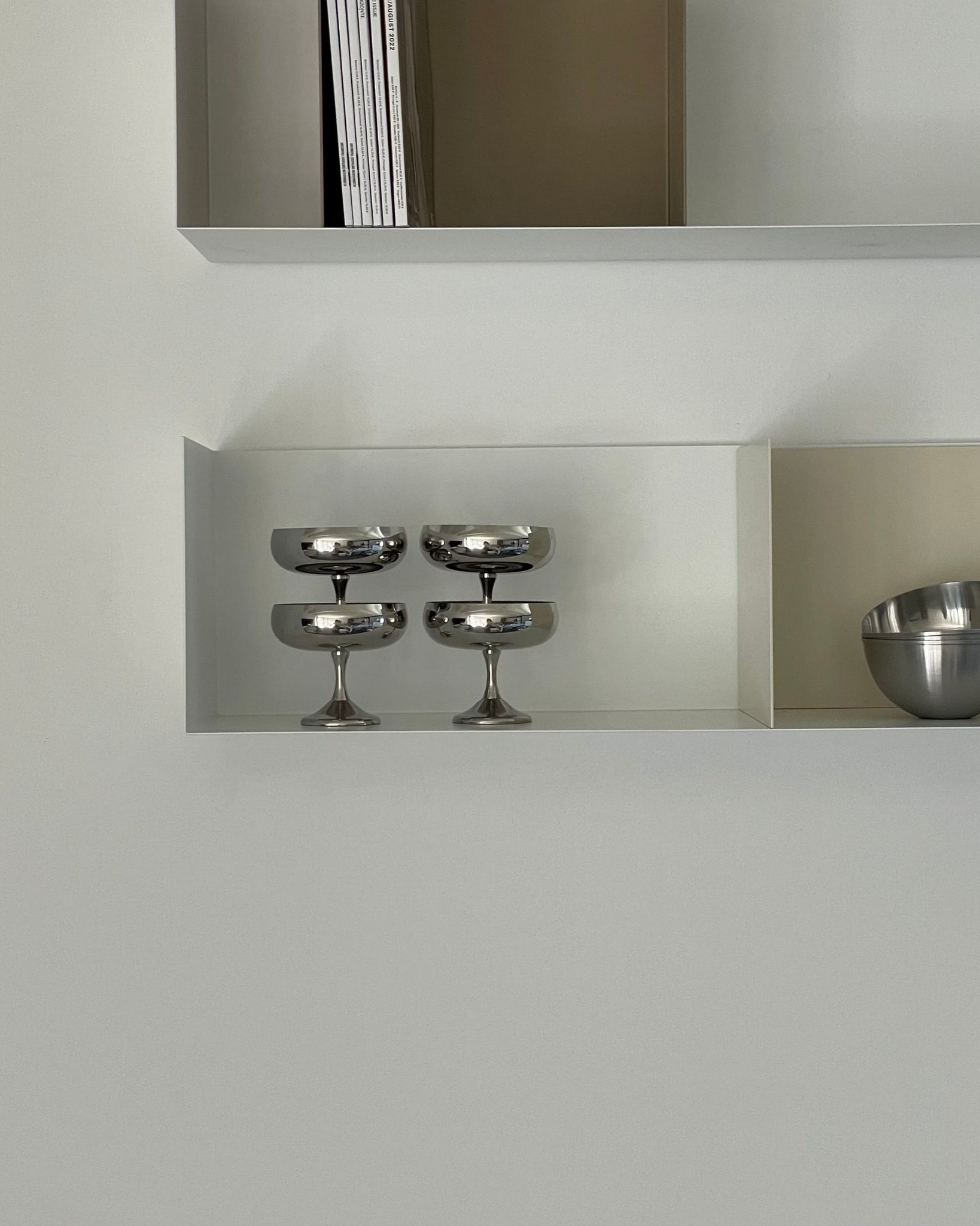 Weißes Wandregal mit Buchstützen in Cremeweiß und Graubeige, dekoriert mit Edelstahl-Schalen und Gläsern