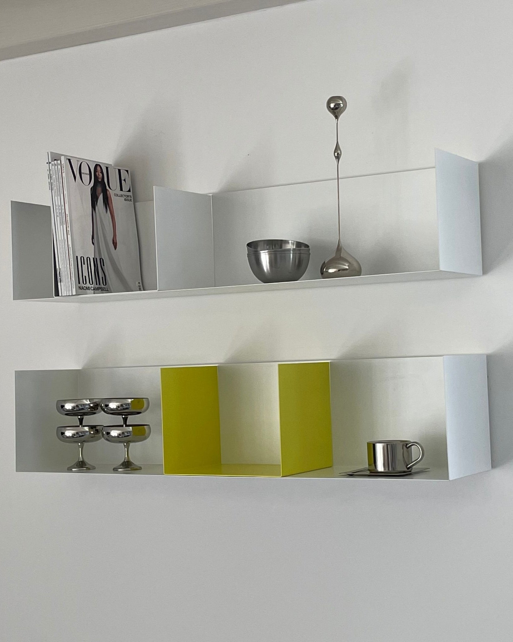 Weißes Wandregal mit gelber Buchstütze, dekoriert mit Edelstahl-Accessoires und Magazinen