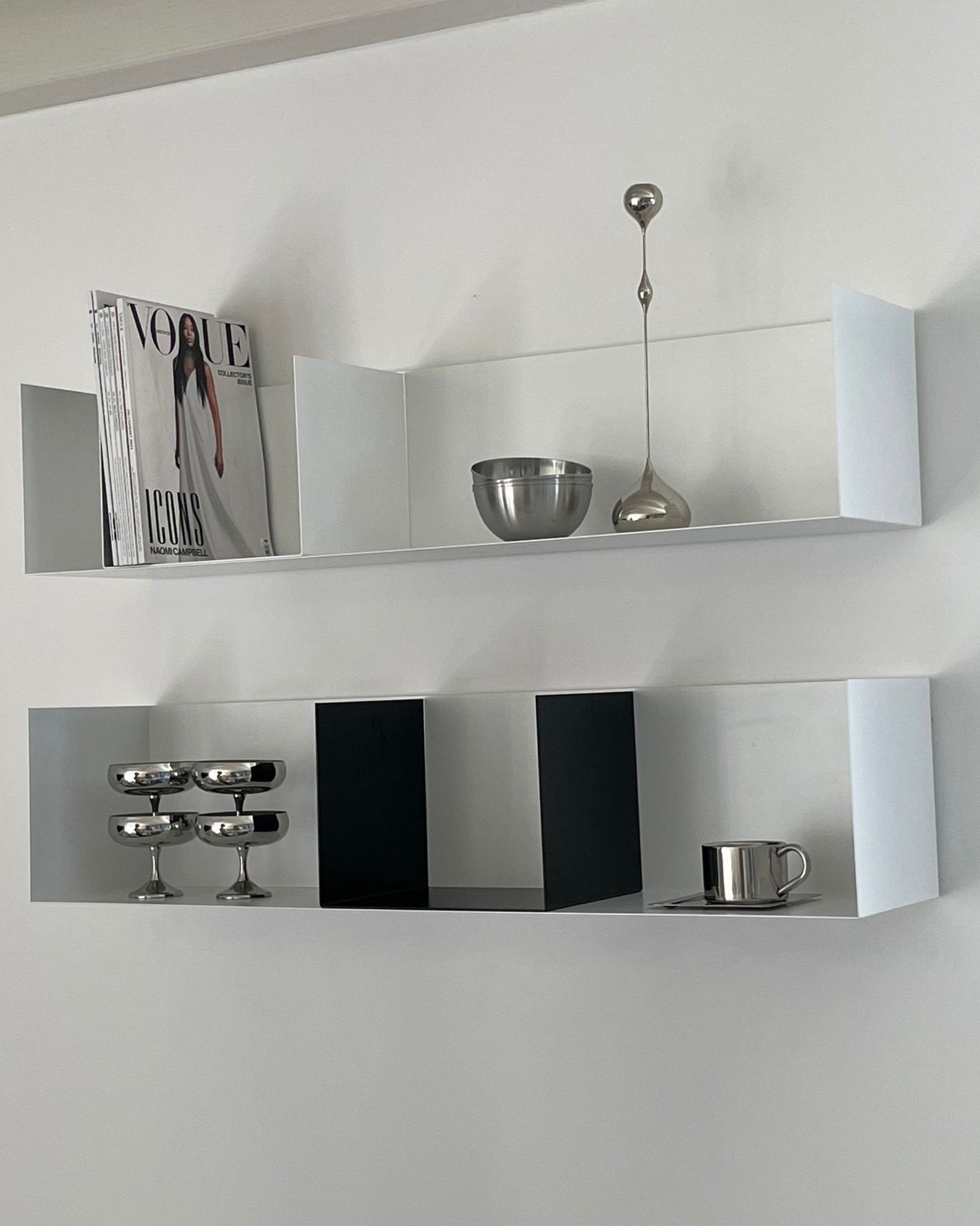 Weißes Wandregal mit schwarzen Buchstützen, dekoriert mit Edelstahl-Accessoires und Magazinen