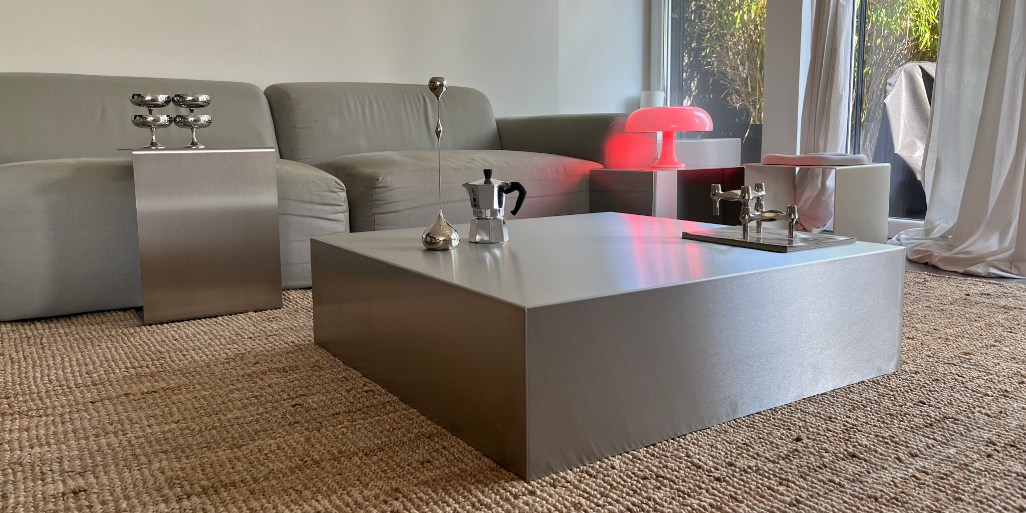 Modernes Wohnzimmer mit großem quadratischen Couchtisch und schlankem Beistelltisch aus gebürstetem Edelstahl, dekoriert mit silbernen Accessoires, vor grauem Sofa und roter Tischlampe