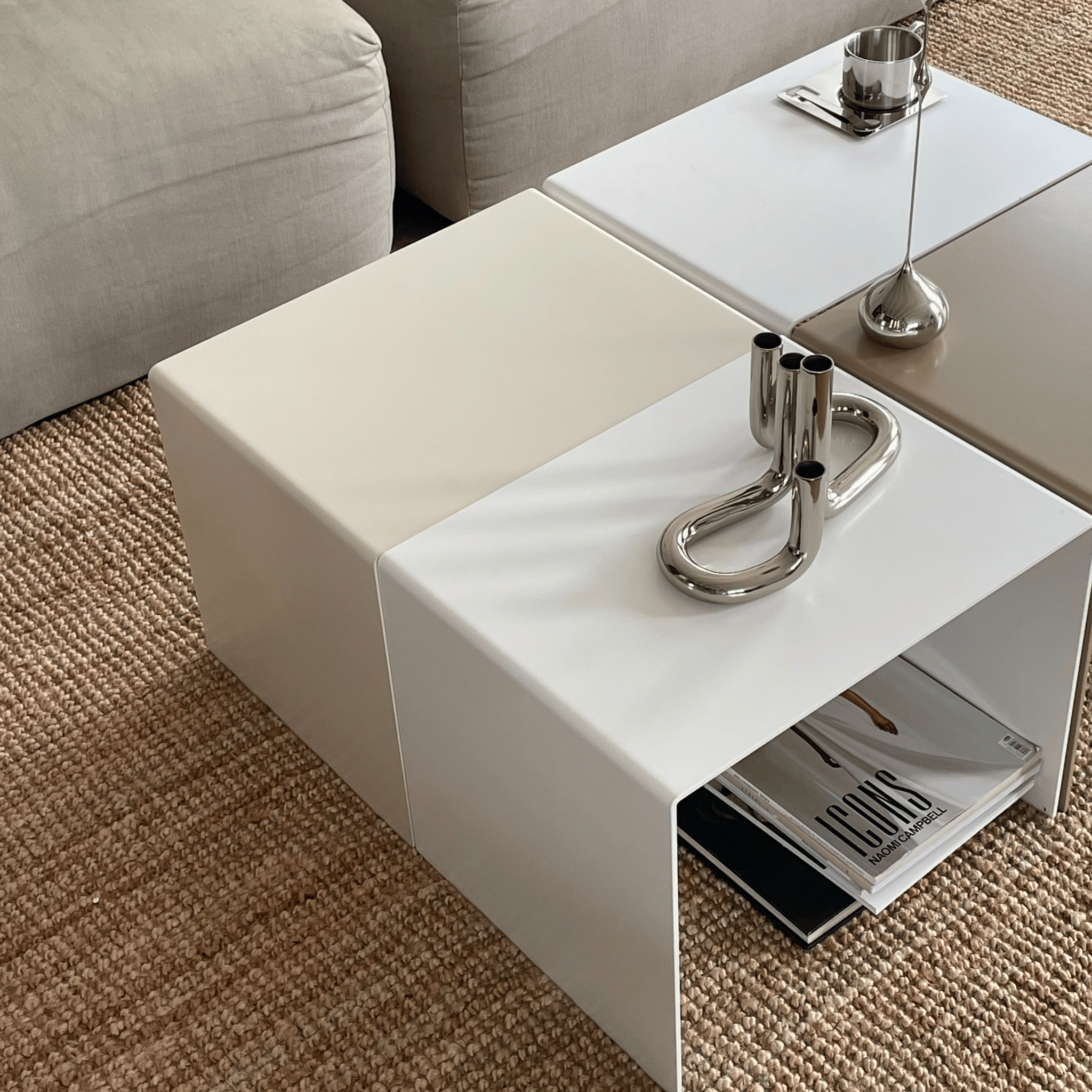 Moderner weißer Beistelltisch im minimalistischen Design, kombiniert mit weiteren Beistelltischen in Beige und Grau, dekoriert mit silbernen Accessoires, auf einem gewebten Teppich vor einem grauen Sofa