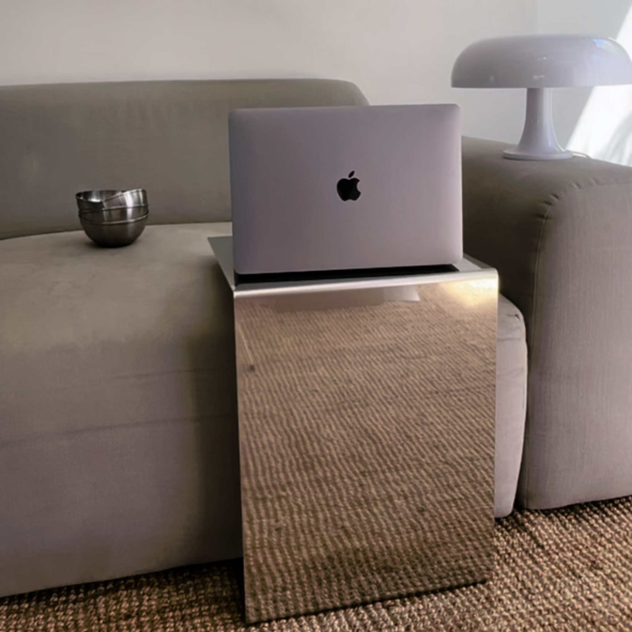 MO INTERIORS spiegelpolierter Beistelltisch mit Laptop, neben einem grauen Sofa und einer modernen Tischlampe, auf einem strukturierten Teppich – minimalistisches und elegantes Interieur.