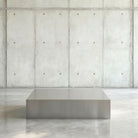 Brushed Q Couchtisch in gebürstetem Edelsthahl auf grauen Betonboden vor grauer Betonwand mit Löchern