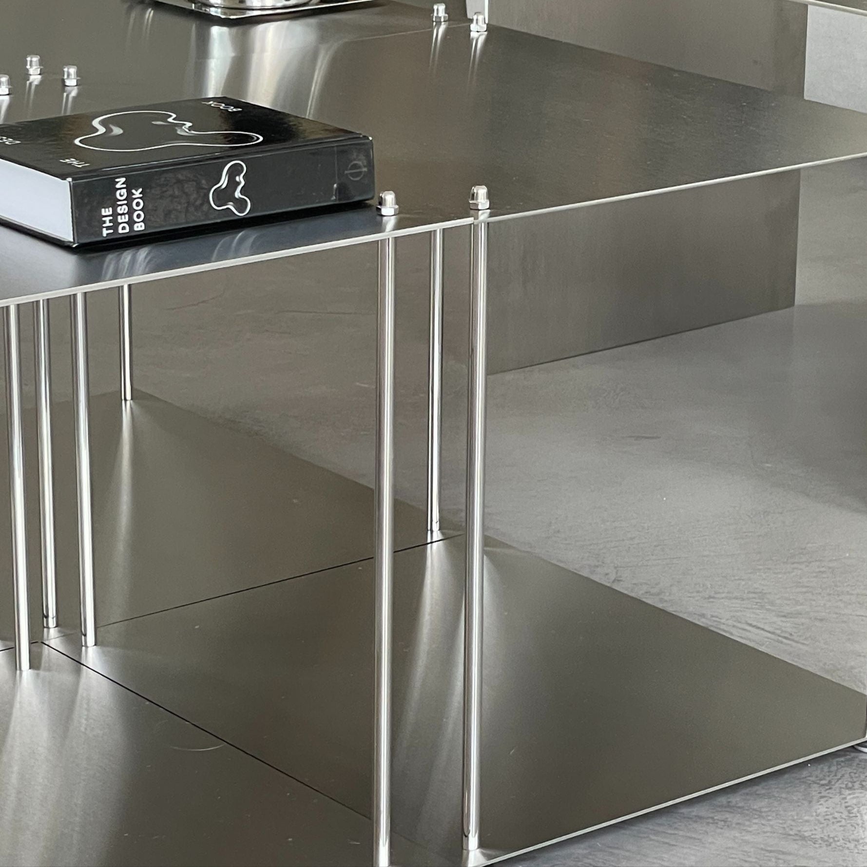 Nahaufnahme eines quadratischen Modulum Couchtisches aus gebürstetem Edelstahl mit zwei Ebenen und sichtbaren Verschraubungen auf glattem Boden im modernen Wohnraum
