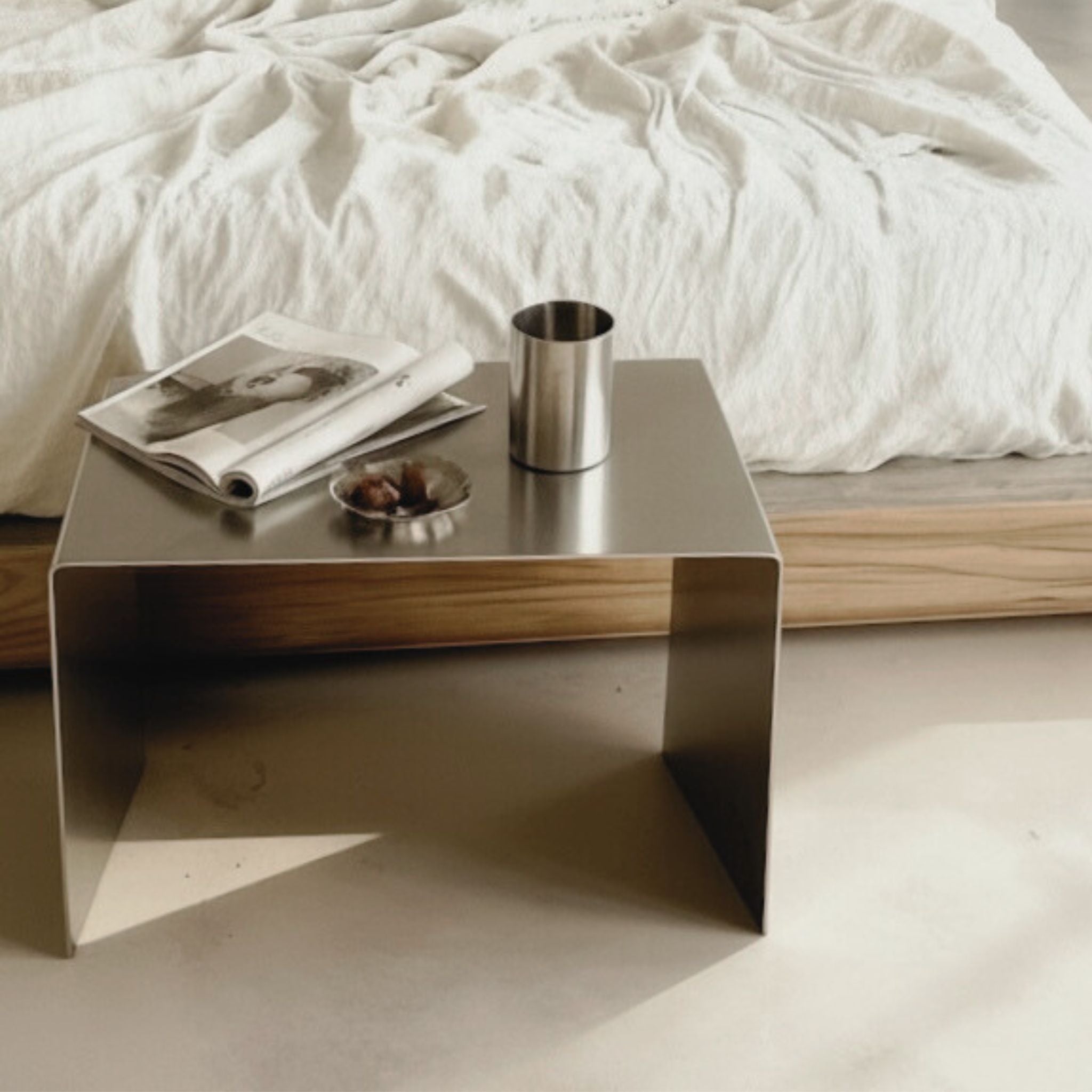 Brushed C Beistelltisch aus gebürstetem Edelstahl als minimalistischer Nachttisch mit Magazin und Becher im Schlafzimmer