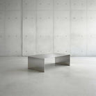 Brushed C Long Couchtisch aus gebürstetem Edelstahl in minimalistischer Studioinszenierung vor Betonwand