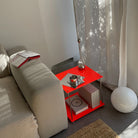 Leuchtroter Design-Beistelltisch aus Edelstahl neben Sofa mit Lampe, Büchern und Vorhang im Sonnenlicht
