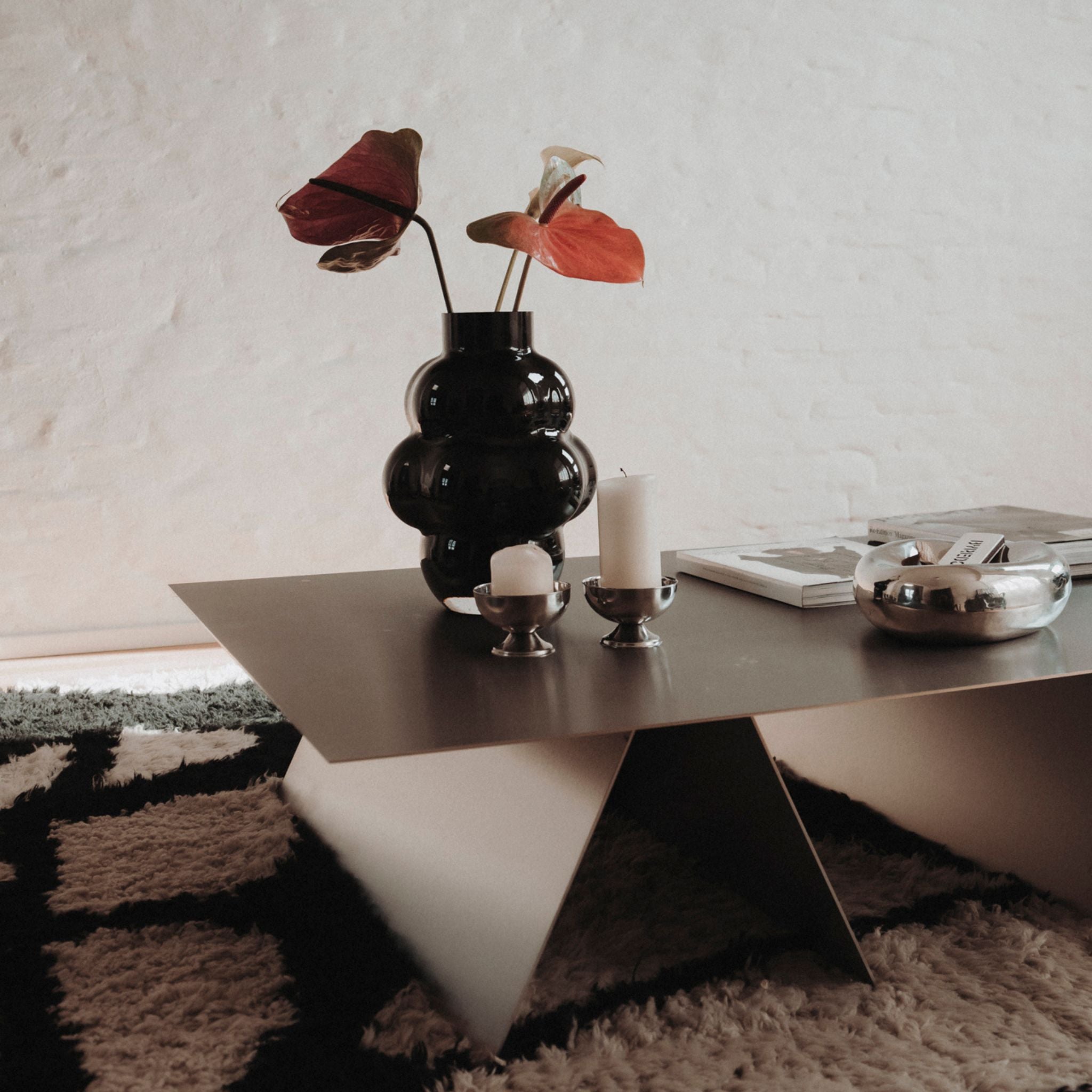 couchtisch aus edelstahl mit vase und kerzen auf designerteppich vor weißer wand in modernem wohnzimmer