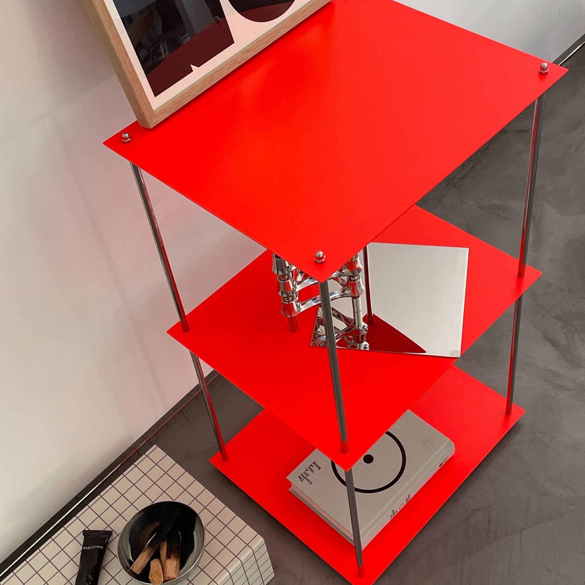 Modulares Designregal MODULUM in Leuchtrot mit drei Böden, dekoriert mit Buch und Kerzenhalter, vor weißer Wand auf Betonboden