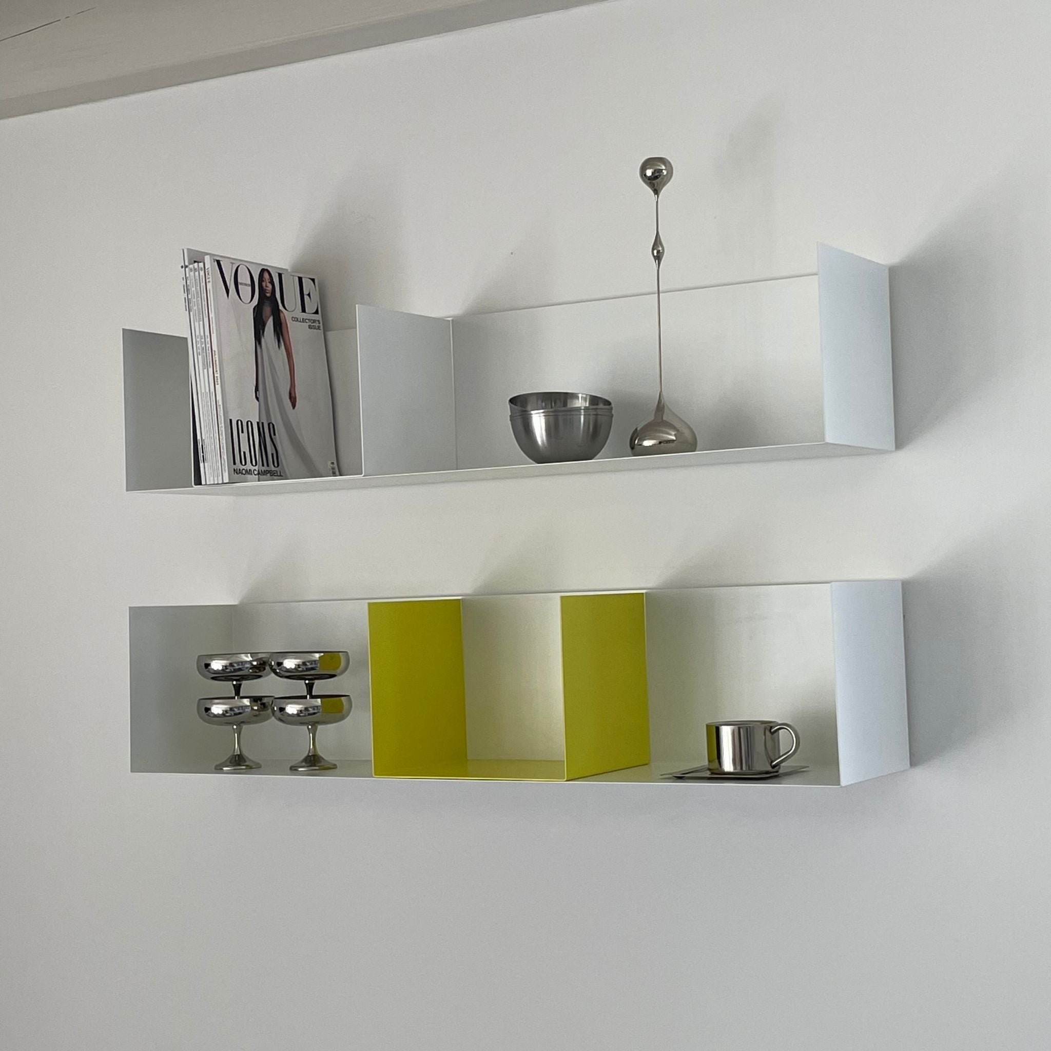 Weißes Wandregal mit gelber Buchstütze, dekoriert mit Edelstahl-Accessoires und Magazinen